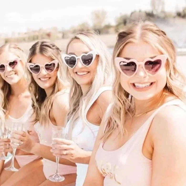 Four women celebrating a bachelorette party outdoors in Destin Florida wearing heart-shaped sunglasses and holding champagne glasses