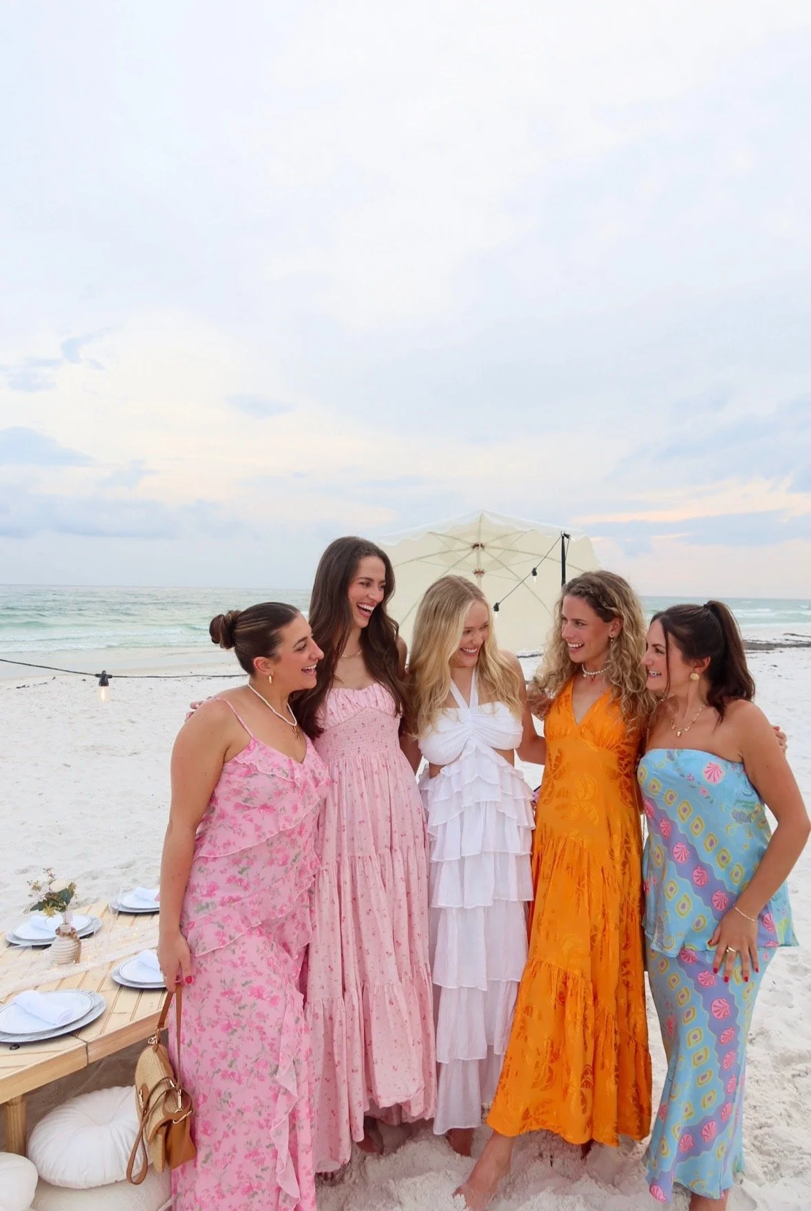 Five women in colorful dresses enjoying a beach gathering under a partly cloudy sky with string lights and a white umbrella.