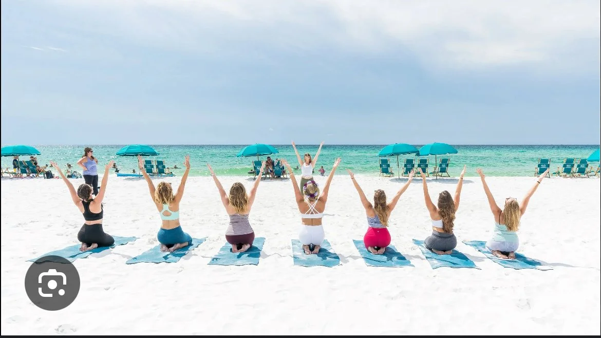 Seven women practicing yoga on a beach with blue umbrellas and chairs, facing the ocean.