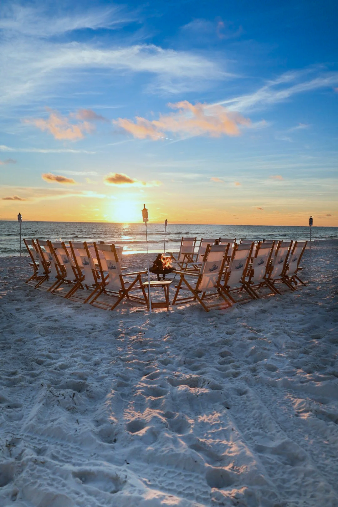 Beach scene at sunset with a circle of chairs around a fire pit, on sandy beach facing the ocean.