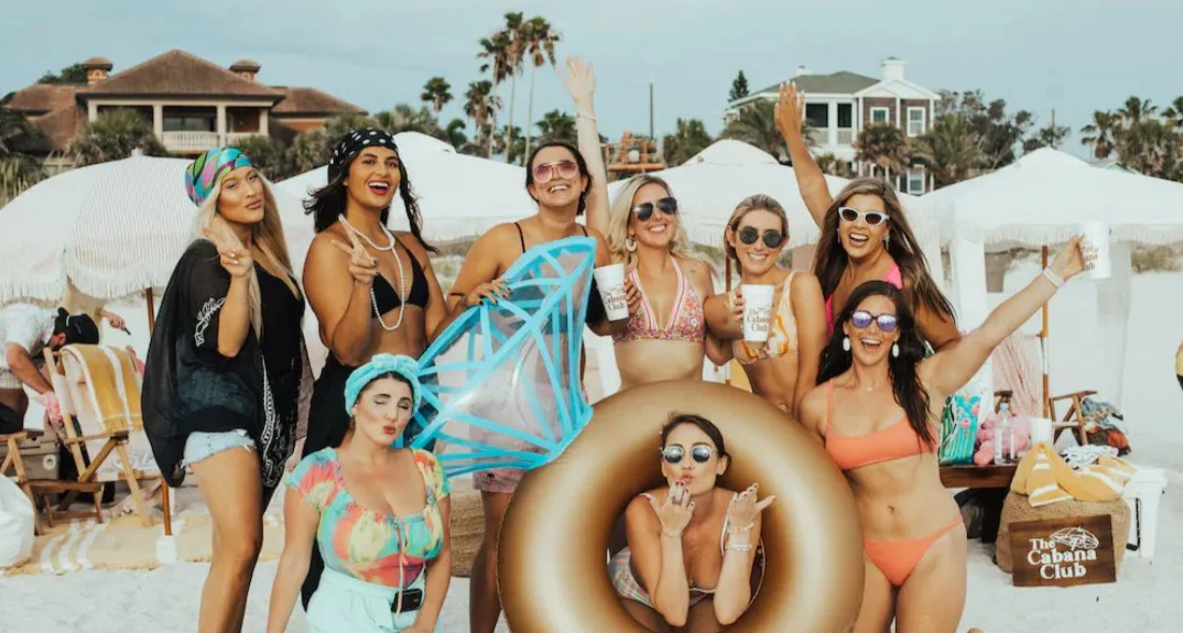Group of smiling women in swimsuits and sunglasses at the beach, some holding drinks, one with a large inflatable ring and a necklace, others waving or making peace signs, with beach umbrellas and houses in the background.