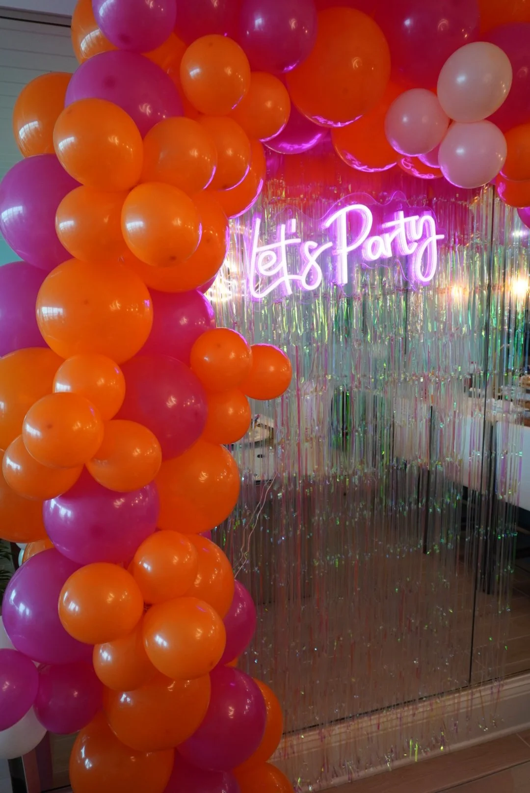 Colorful party decoration with pink, purple, orange, and white balloons arranged around a neon sign that reads "Let's Party" and shiny metallic streamers in the background.