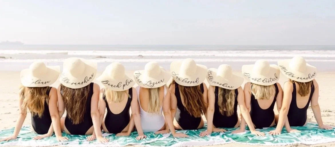 Group of women sitting on a beach with their backs facing the camera, all wearing large white sun hats with cursive writing, black bathing suits, and sitting on a towel near the ocean.