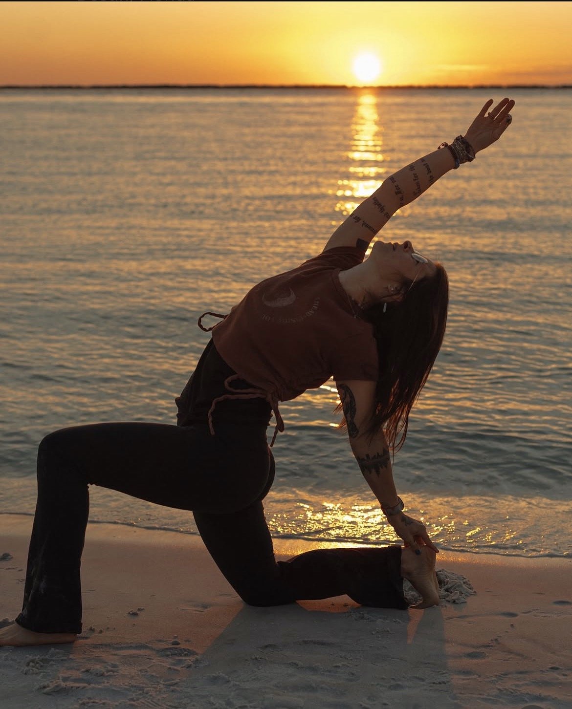 A woman practicing yoga on the beach at sunset, in a lunge pose with one arm reaching overhead and the other hand on her foot, with the ocean and setting sun in the background.