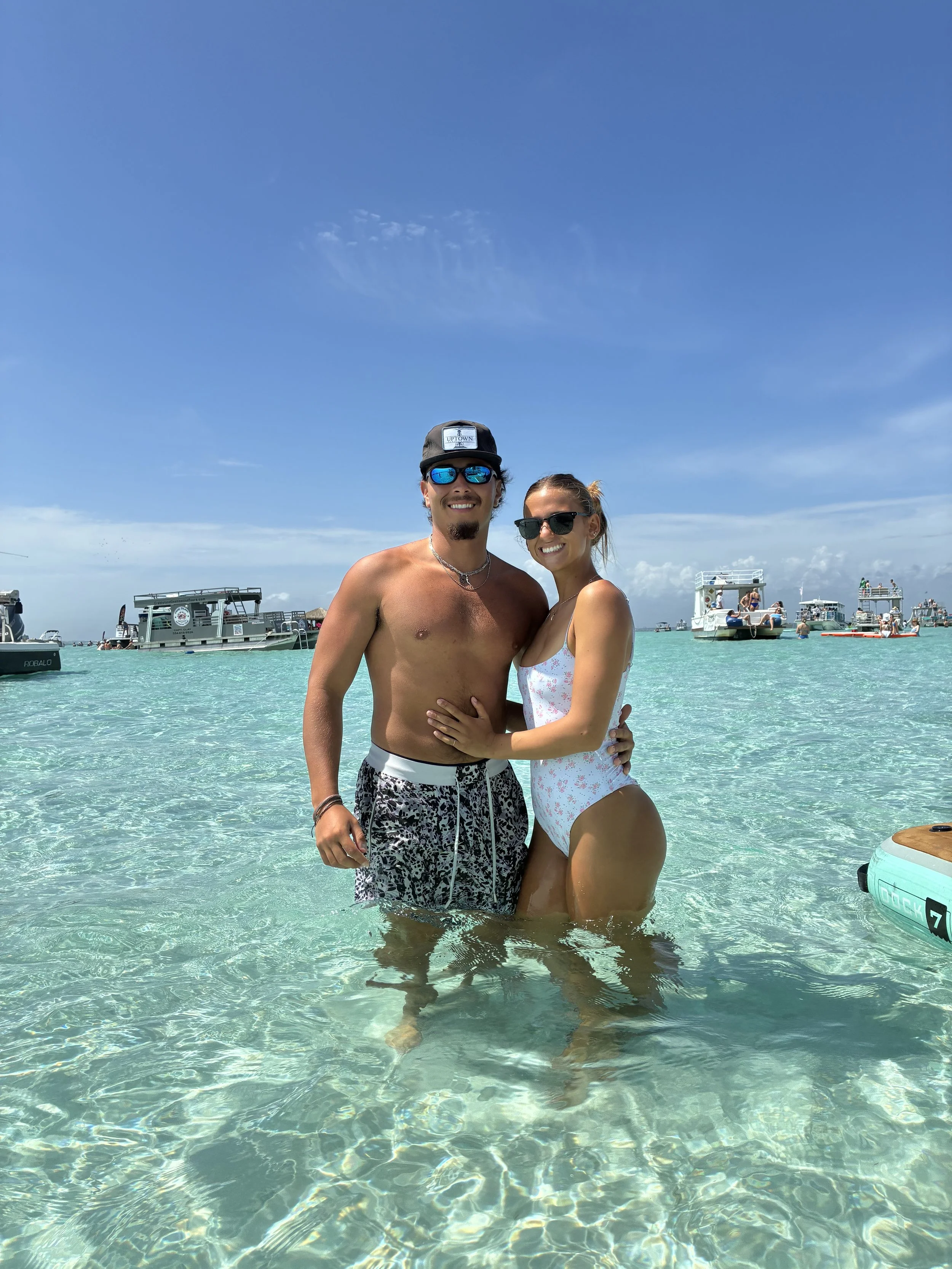 A couple standing in clear, shallow ocean water at the beach, with boats and a clear blue sky in the background. The man is shirtless wearing patterned swim trunks and sunglasses, and the woman is in a white swimsuit with pink floral pattern, also wearing sunglasses.
