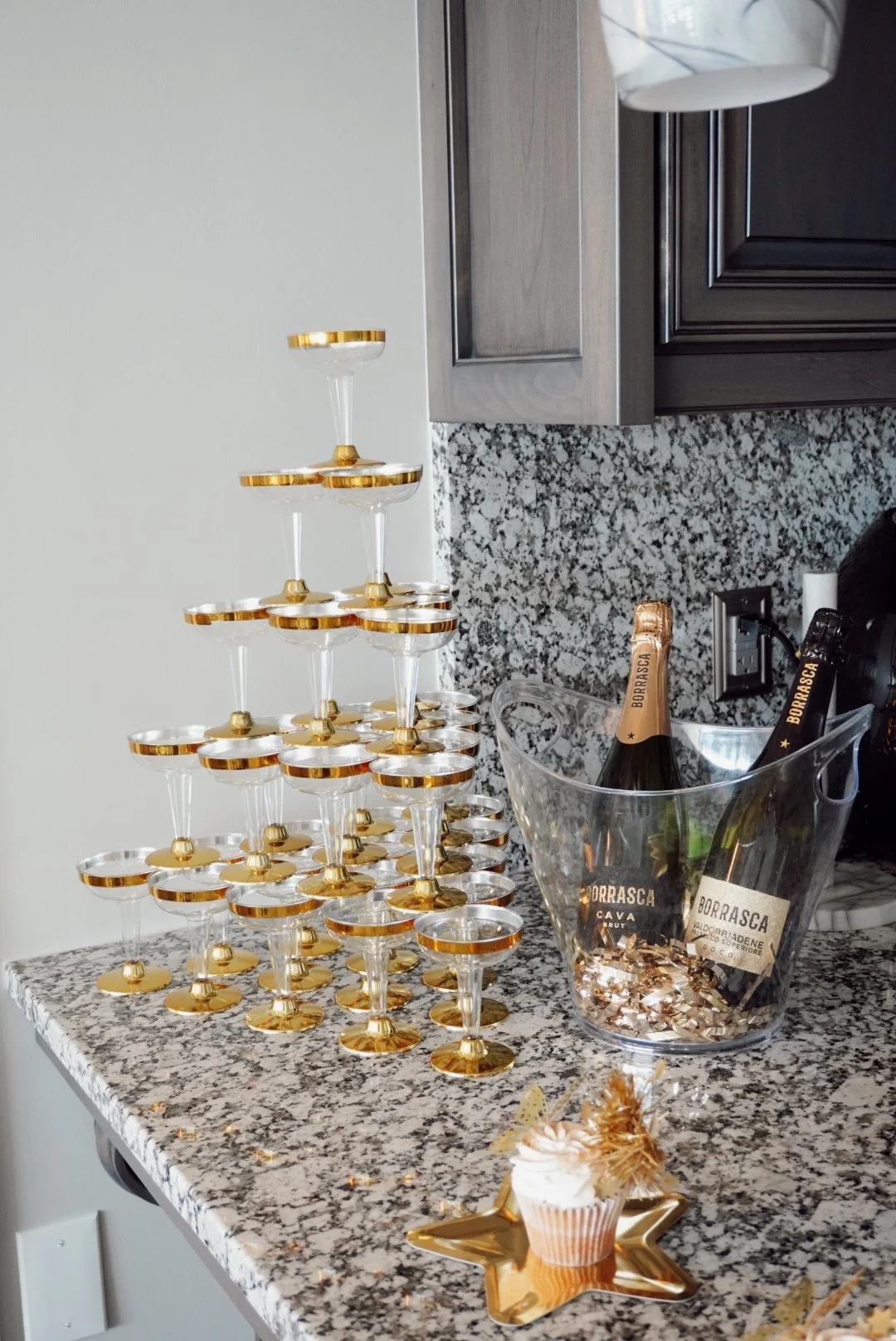 A pyramid of empty champagne glasses with gold rims, bottles of Borrasca Cava in a clear ice bucket, and a small cupcake on a gold star-shaped plate on a granite kitchen countertop.