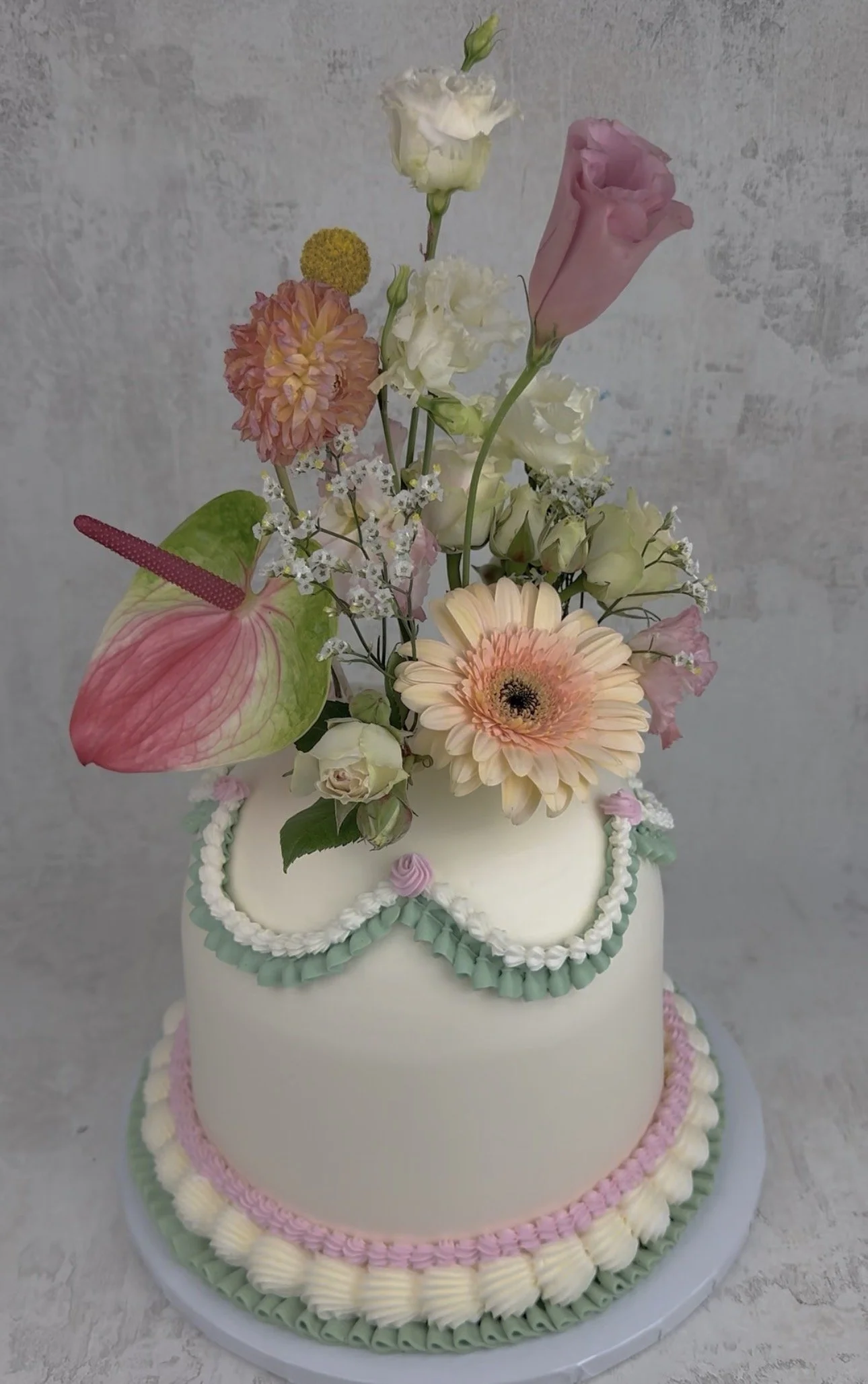 A two-tiered cake with white frosting, decorated with pastel green, pink, and white piped icing around the edges. The top of the cake has a floral arrangement including pink eustoma, peach gerbera daisy, white roses, baby's breath, and an anthurium w