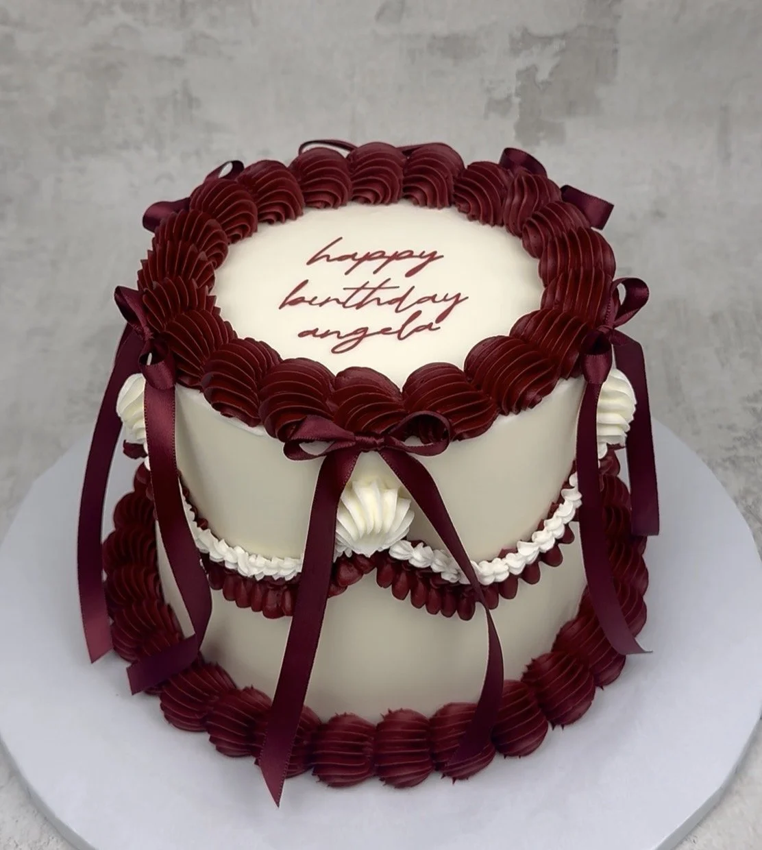 A two-tiered birthday cake decorated with white and burgundy frosting, burgundy ribbon bows, and piped frosting patterns. The top of the cake has a message that reads 'happy birthday angela' in red script.