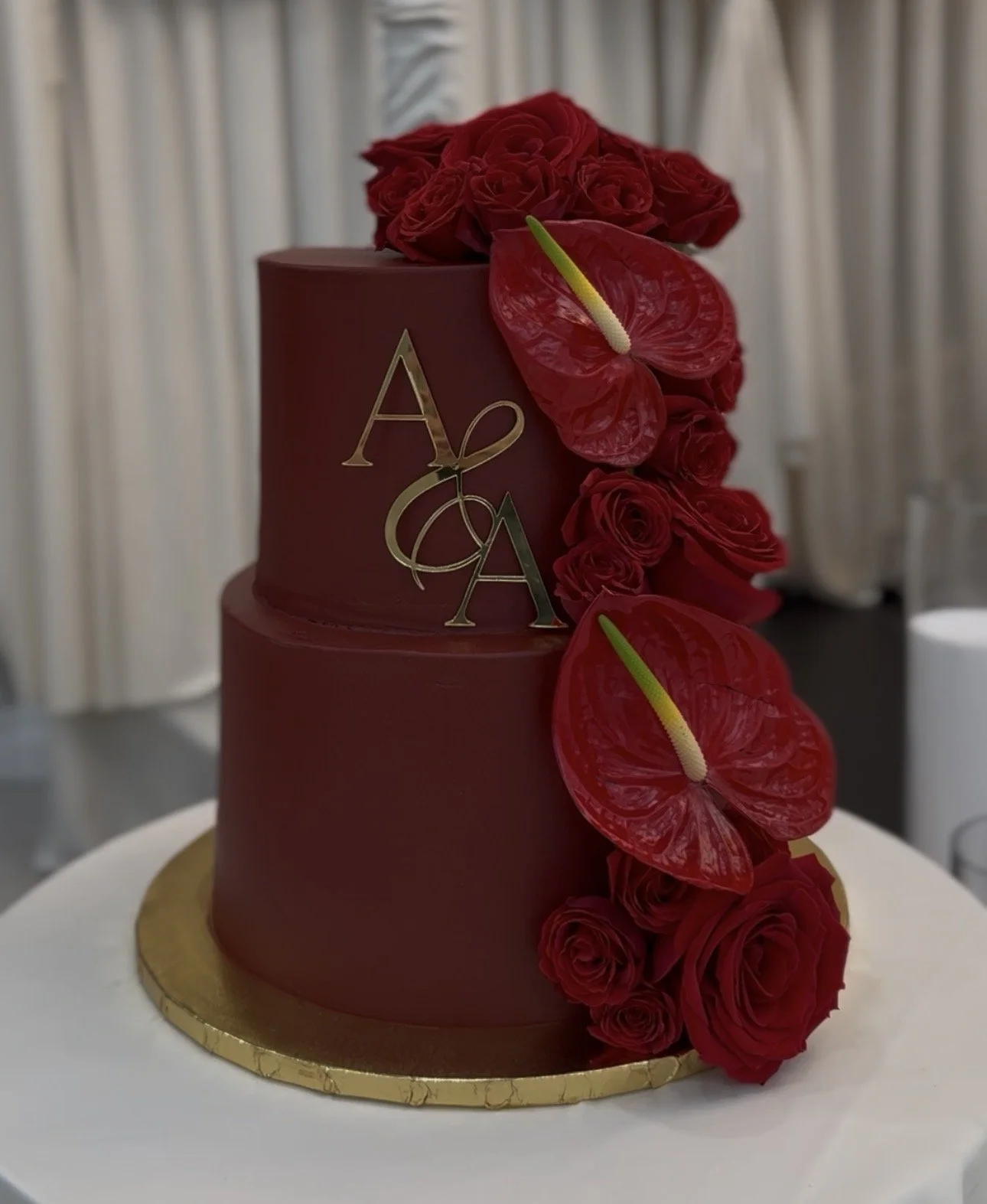 A two-tier red wedding cake decorated with red roses and anthuriums, with gold initials 'A & A' on the front, sitting on a white table with a gold-edged base.