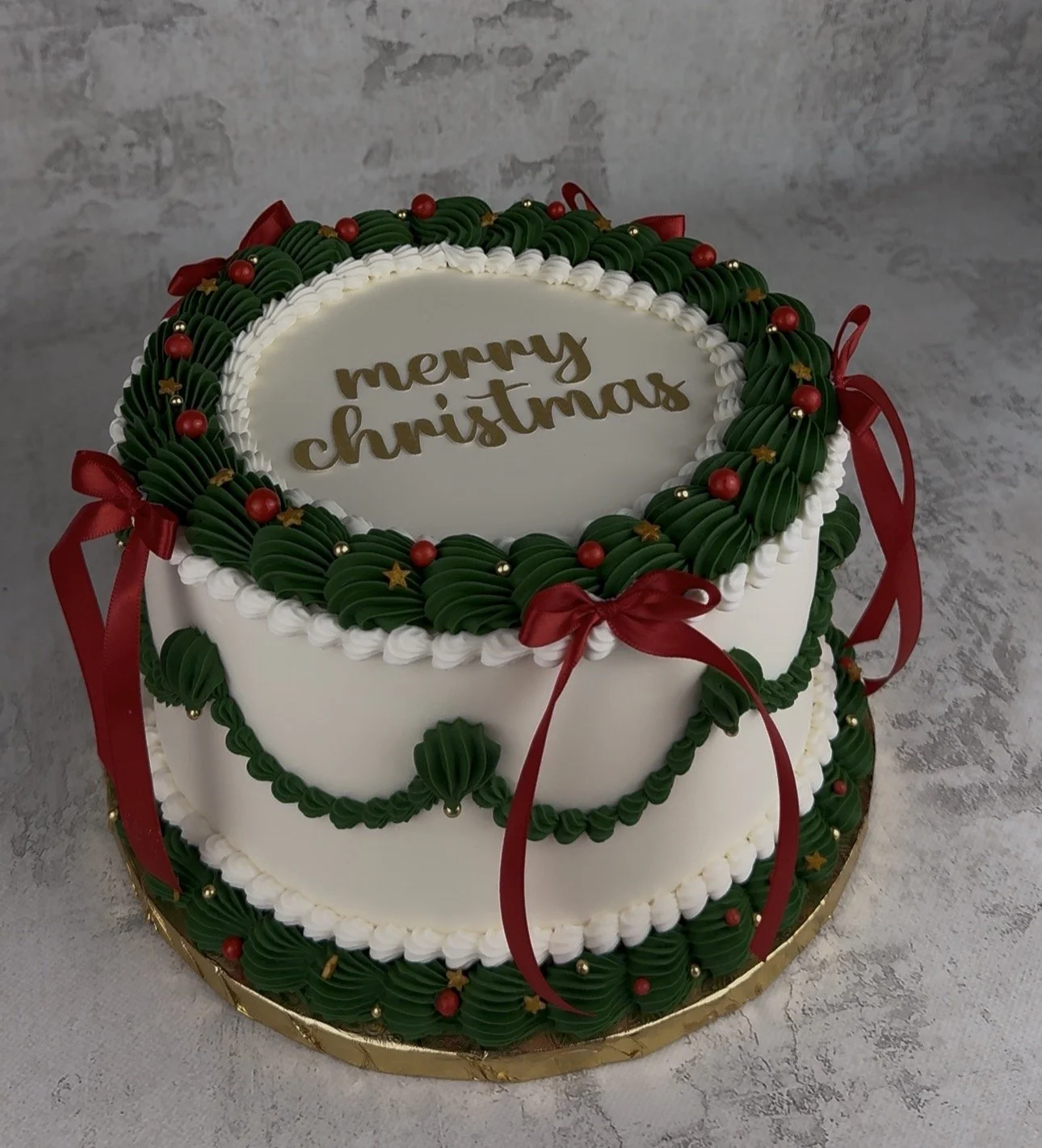A Christmas cake decorated with green frosting, red ribbons, gold beads, and gold star accents, with the message 'Merry Christmas' written on top in gold lettering.