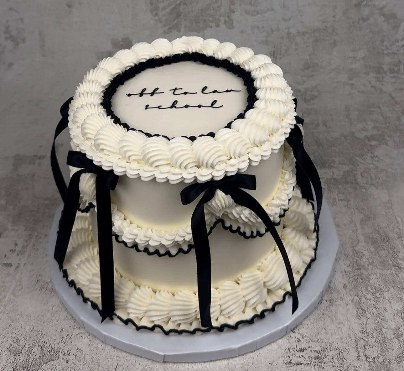 A two-tier white cake decorated with black ribbons and piping, with a message written on top that says "off to law school."