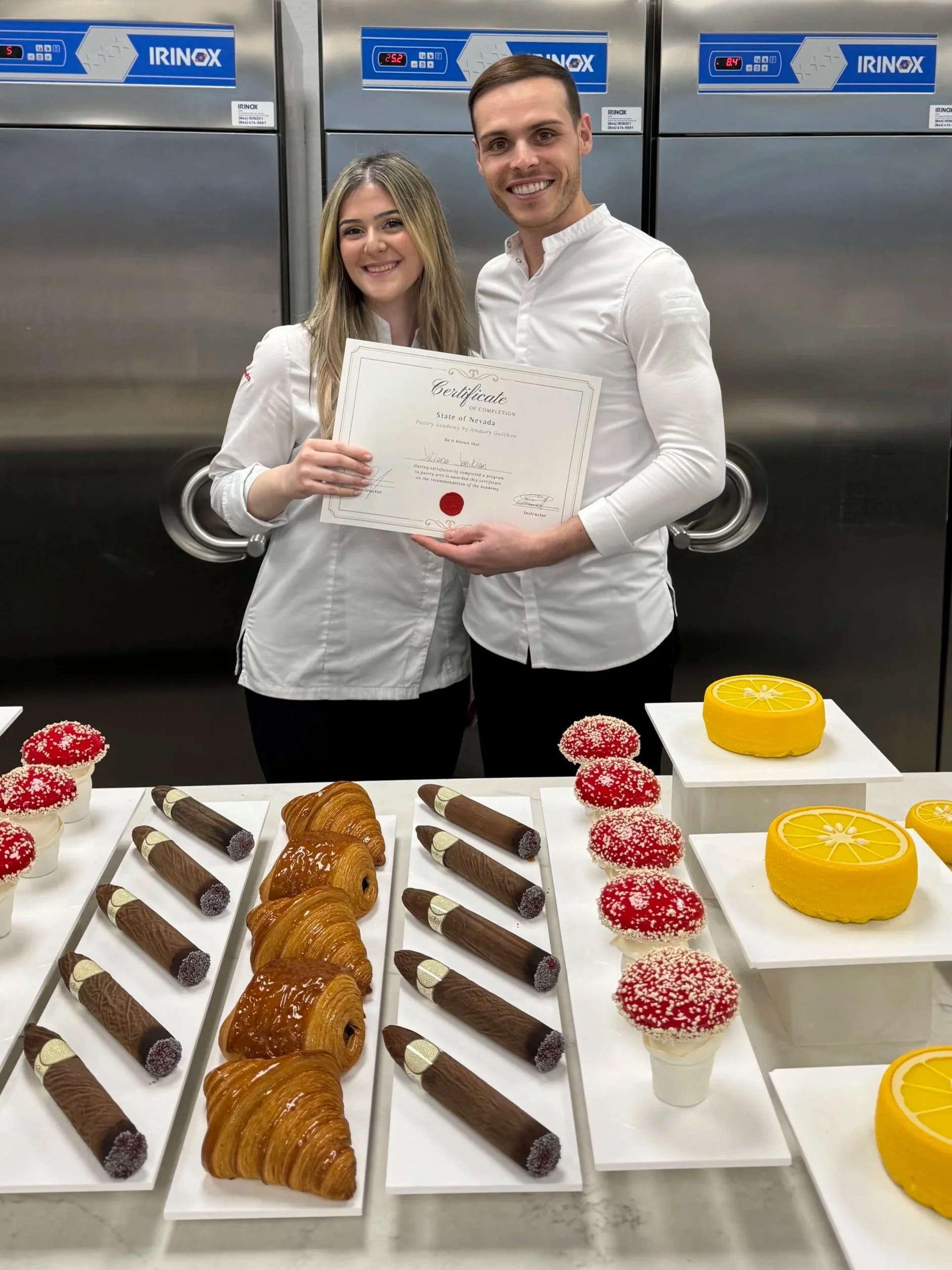 A man and a woman smiling and holding a certificate in a commercial kitchen with stainless steel appliances in the background. On the table in front of them are pastries, including croissants, chocolate rolls, cupcakes with red and white sprinkles, and lemon cakes.