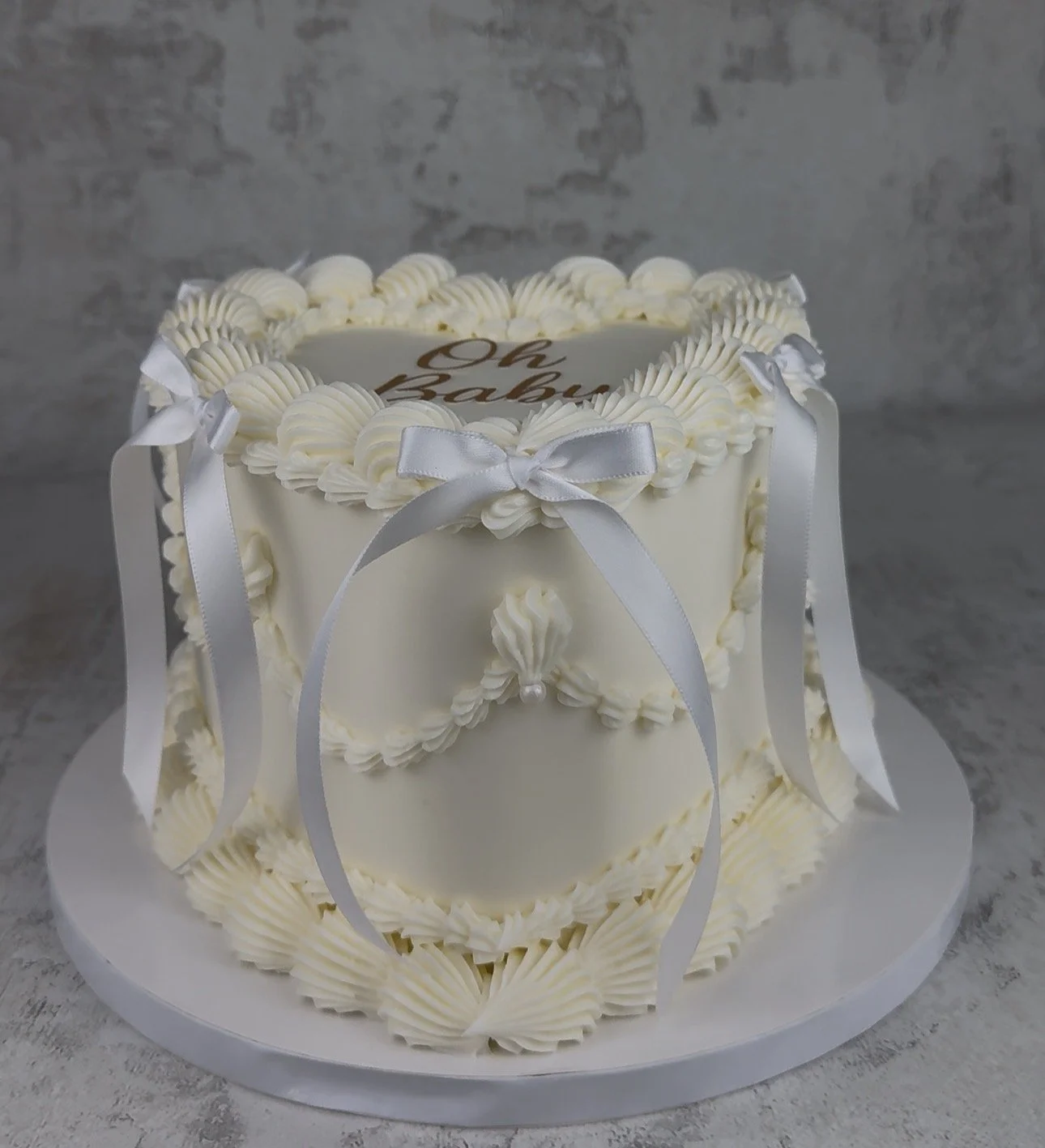 A decorated white cake with piped frosting, white ribbons, and a message on top that reads 'Oh Baby'.