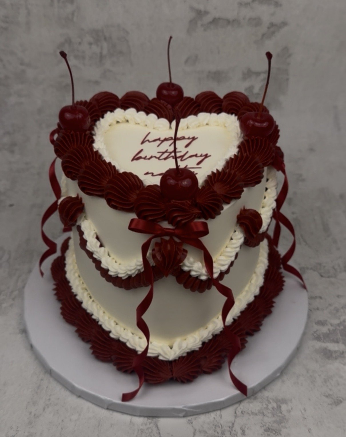 Two-layer heart-shaped birthday cake with white icing, red cherry decorations, and red ribbons, featuring a message written in red on the top heart.
