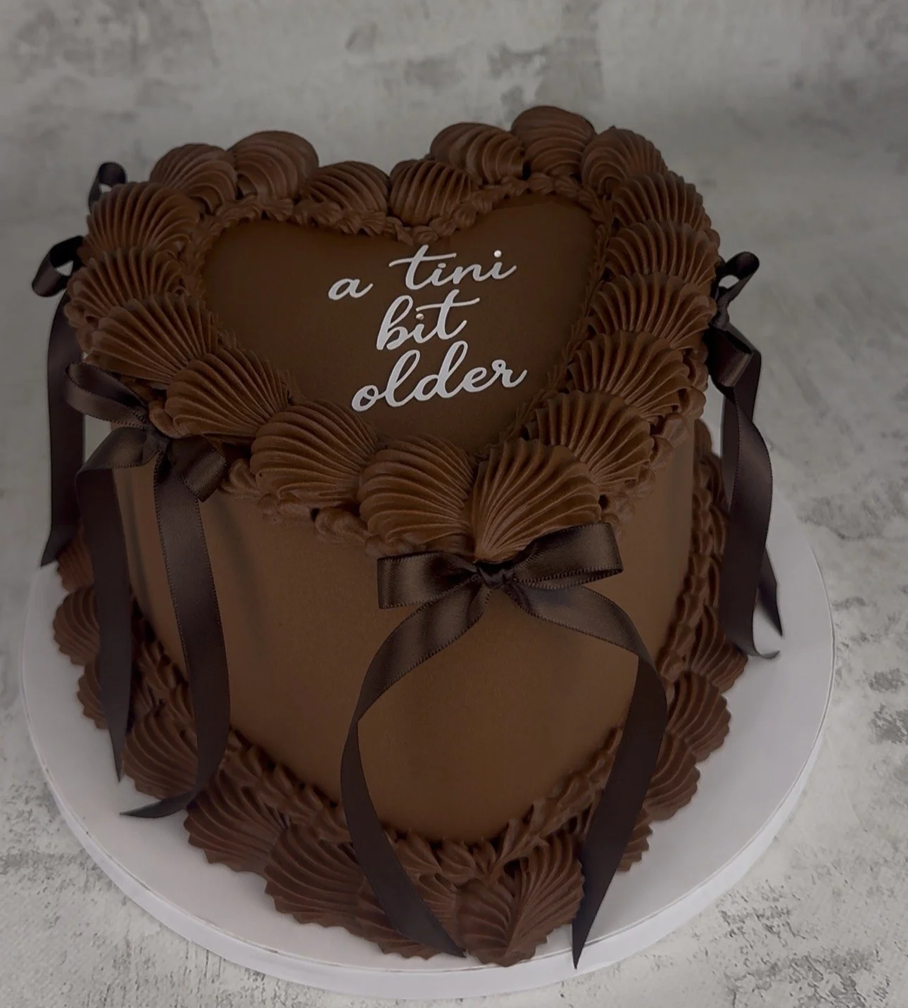 Chocolate heart-shaped cake decorated with piped chocolate cream and ribbon bows, with a message that reads 'a tiny bit older' on top.