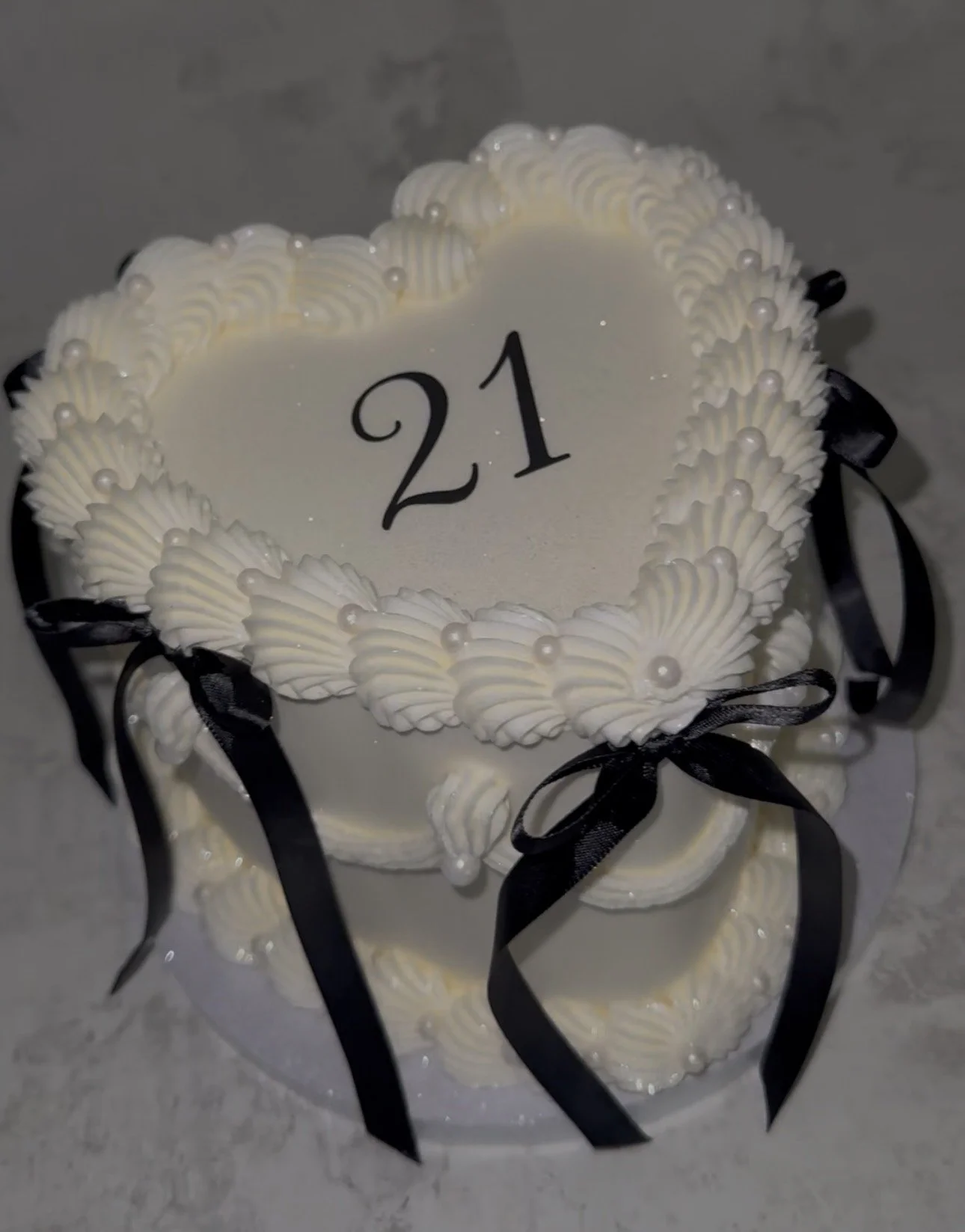 White heart-shaped cake decorated with cream swirls, pearls, and black ribbon bows, with the number 21 on top, likely celebrating a 21st birthday.