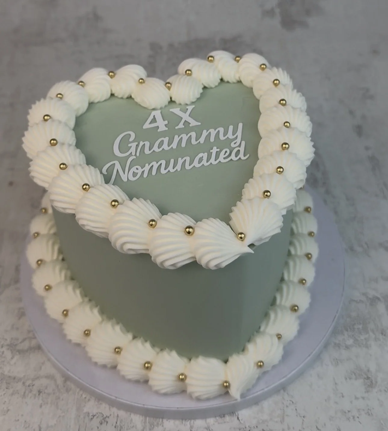 A heart-shaped cake with white frosting decoration and gold ball accents. The cake has a green plaque on the top with the text "4X Granny Nominated."