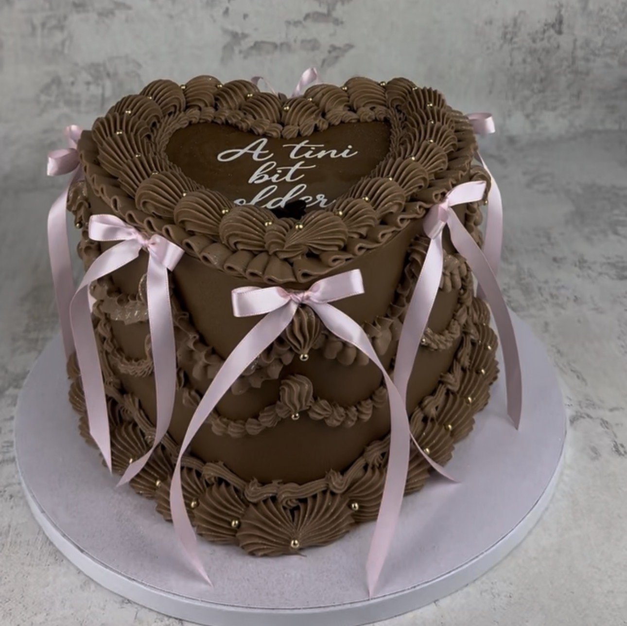 A heart-shaped chocolate cake decorated with pink ribbons and intricate piped icing details, with a message written on top.