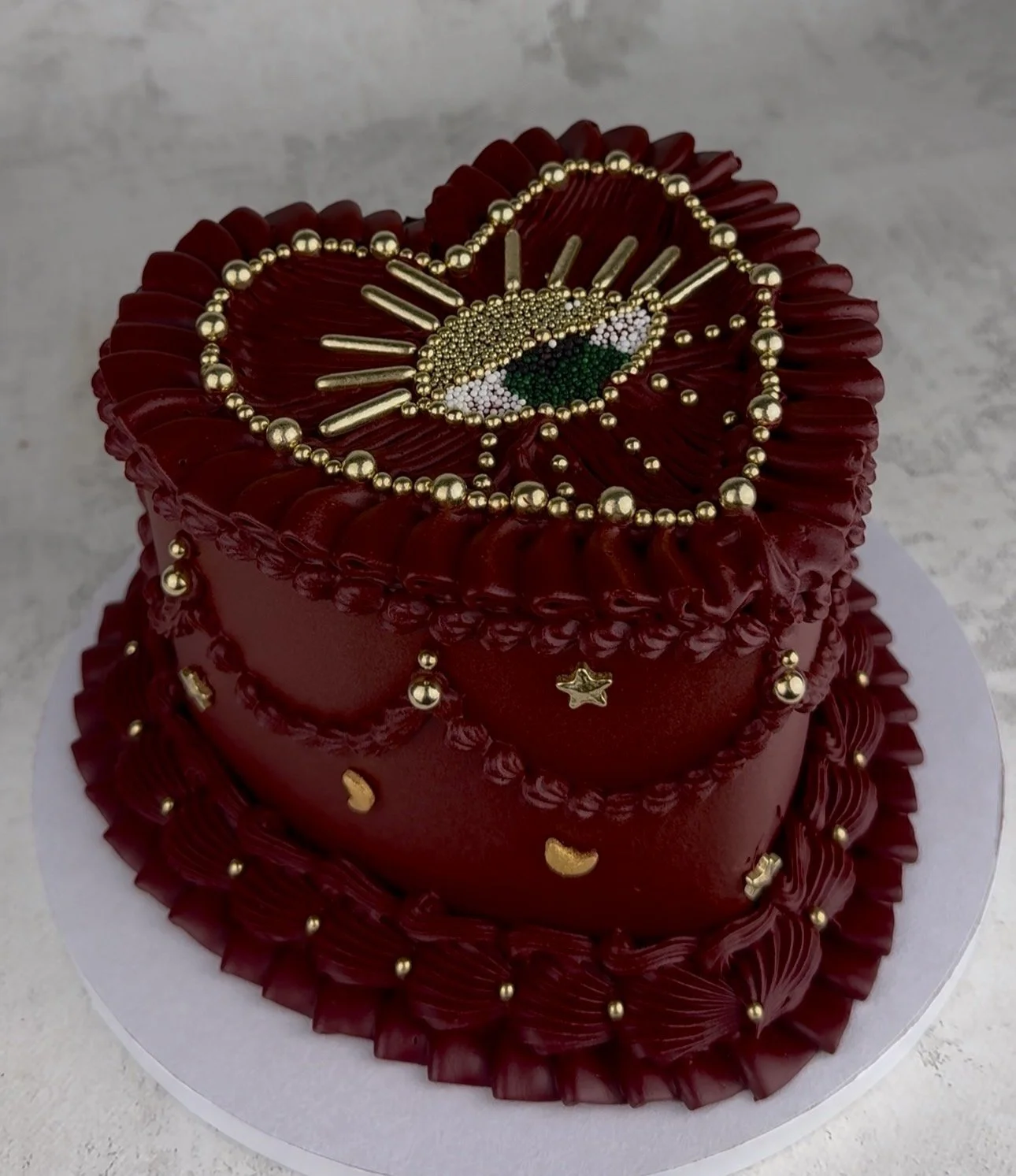 Heart-shaped red cake decorated with gold beads, stars, and crescent moon shapes; topped with a gold eye and bead design.