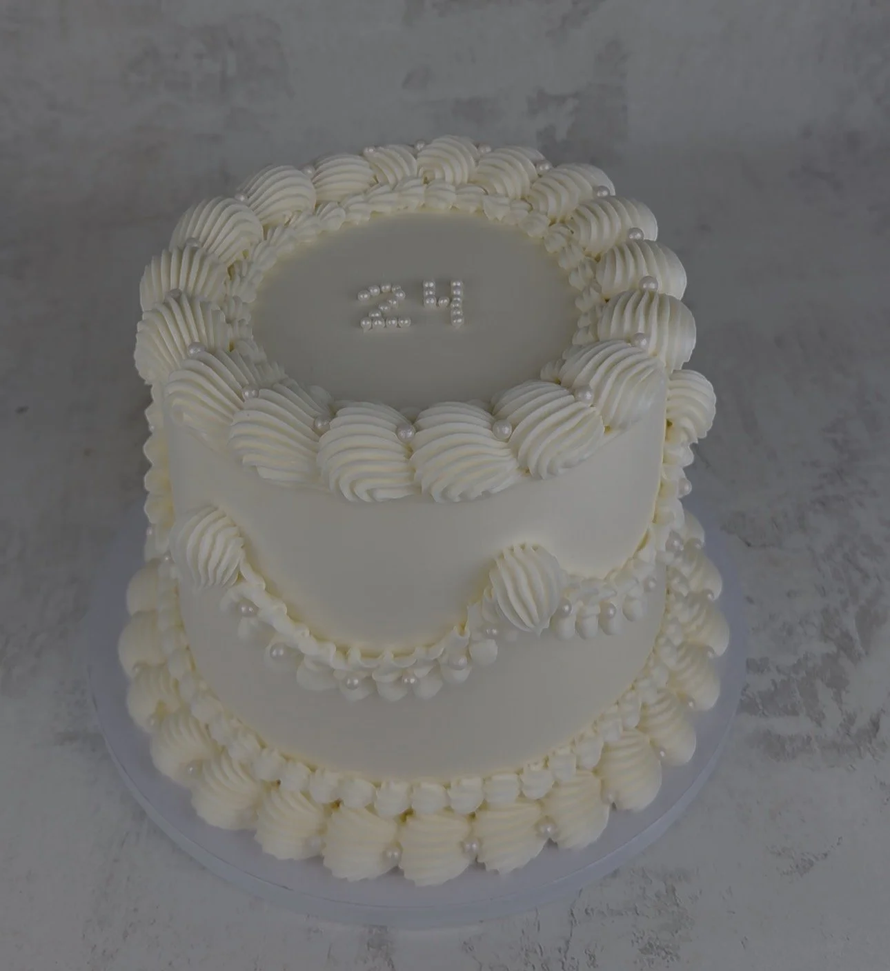 Three-tier white birthday cake decorated with cream swirls and edible pearls, with a silver inside a circle on top displaying the number 24.