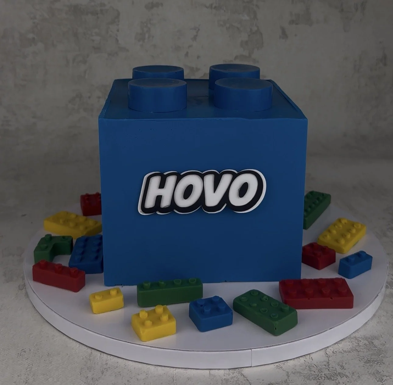 A cake shaped like a blue LEGO brick with the word 'HOVO' on it, surrounded by colorful small LEGO bricks on a white circular platform.