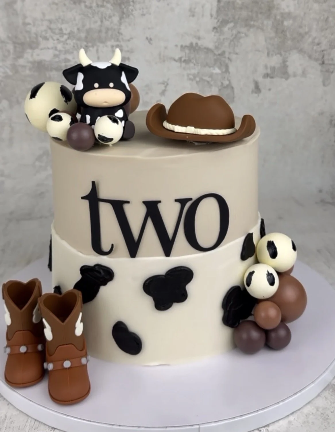Two-tiered cow-themed birthday cake decorated with cow print candies, a cow figurine, a cowboy hat, and small cowboy boots, with the word "two" on the front.