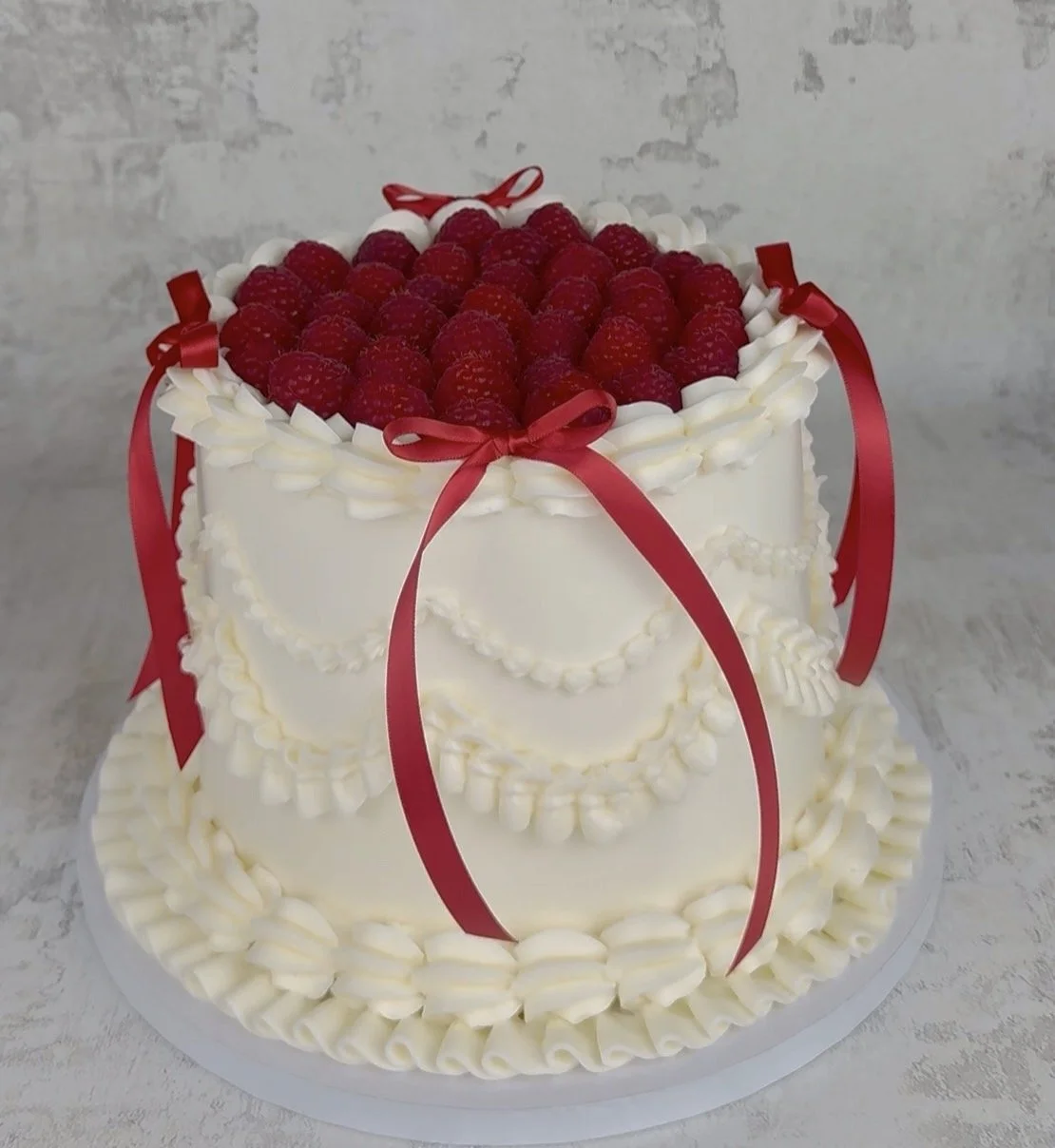 Two-tier white cake decorated with red ribbons, piped white frosting, and topped with raspberries.