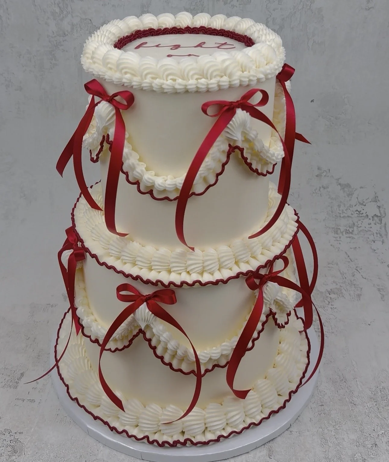 Three-tier white cake decorated with red ribbons, red piping, and white whipped cream border details. The top of the cake has a round border with the word "fruit" written in red.