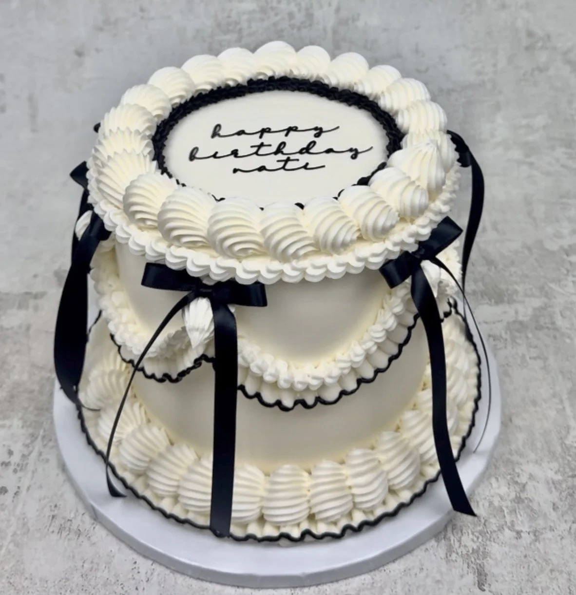 Three-tier white birthday cake with black ribbon decorations and piped white frosting, featuring a message on top that says 'happy birthday nate'
