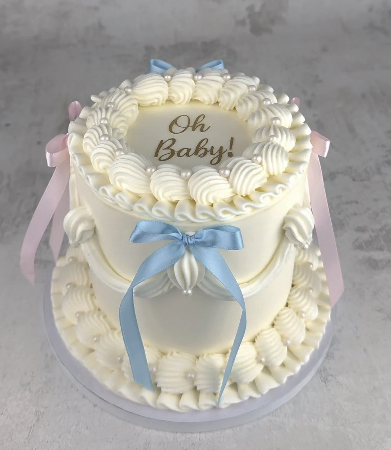 Two-tiered white baby shower cake decorated with cream swirls, pearl-like accents, and pink and blue ribbons. The top of the cake has a circular message that reads 'Oh Baby!' in gold lettering.
