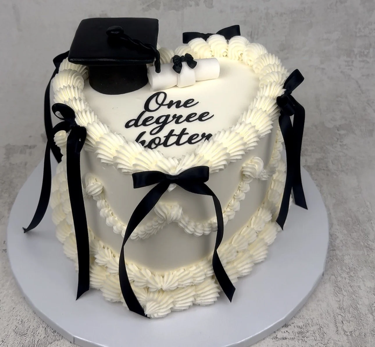 Heart-shaped graduation cake with white frosting, black ribbon decorations, a graduation cap topper, a diploma, and the words "One degree hotter" written on it.