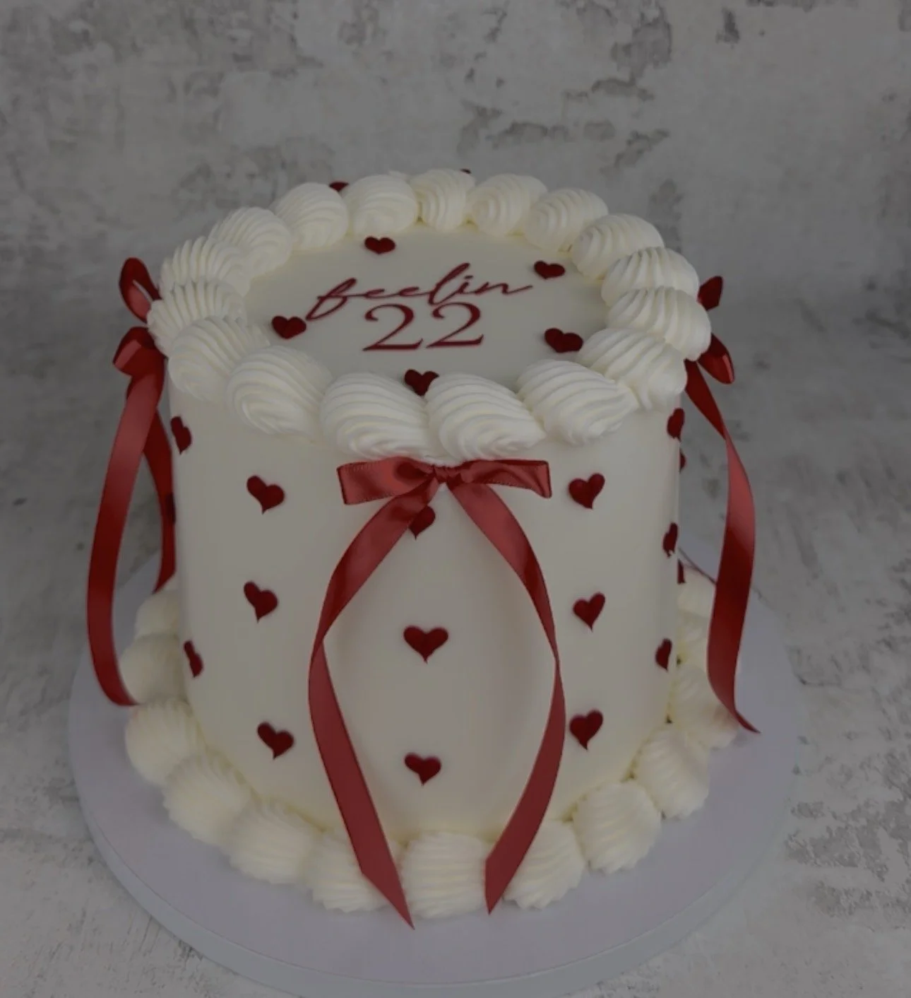 A round, white cake decorated with red hearts, white frosting, and red ribbons. The top has a message written in red that reads "feelin 22" and the cake is set against a gray background.