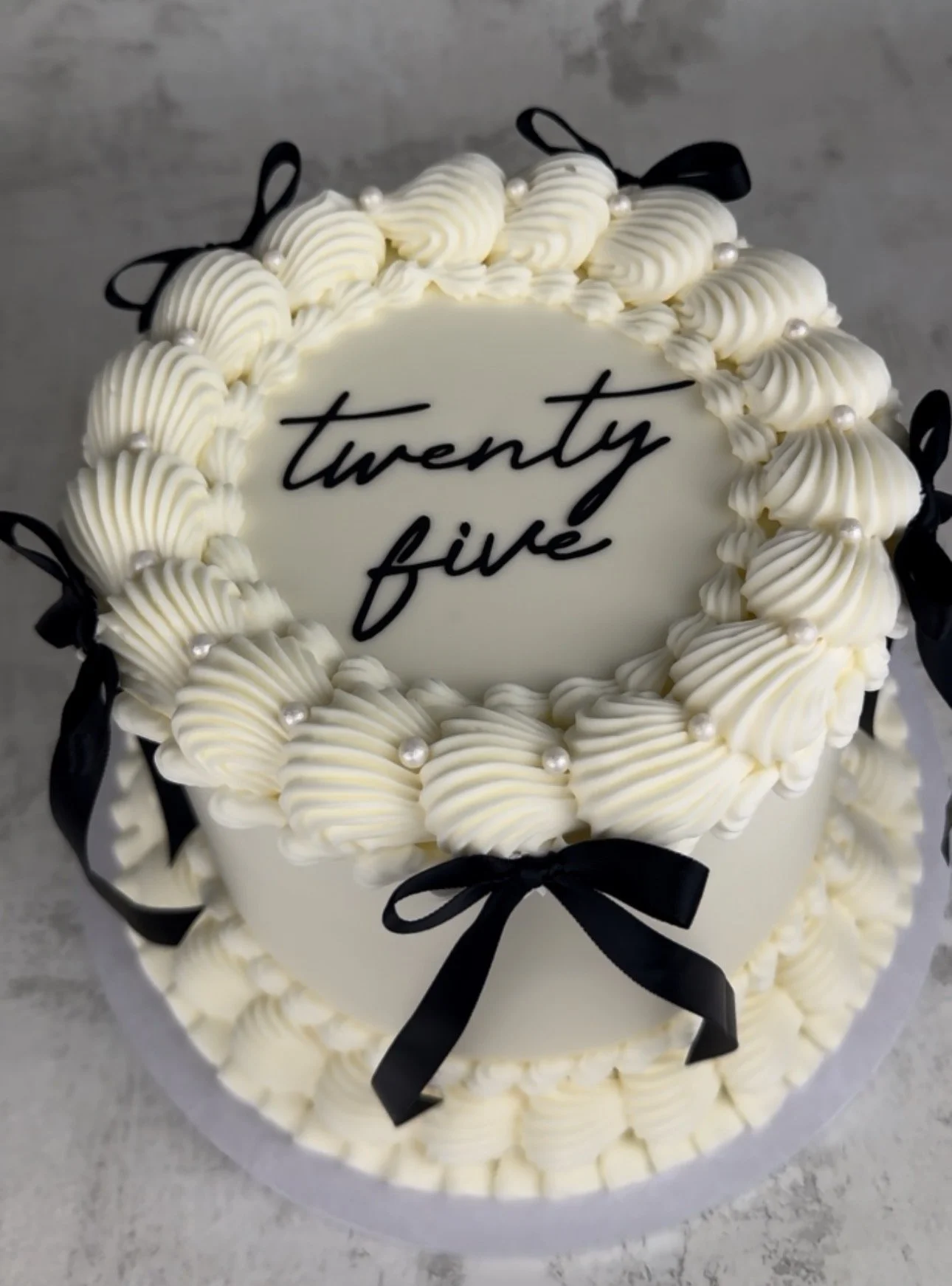 Layered cream-colored birthday cake with white frosting, black ribbons, small pearls around the top, and a white rectangular plaque with the words 'twenty five' written in black.