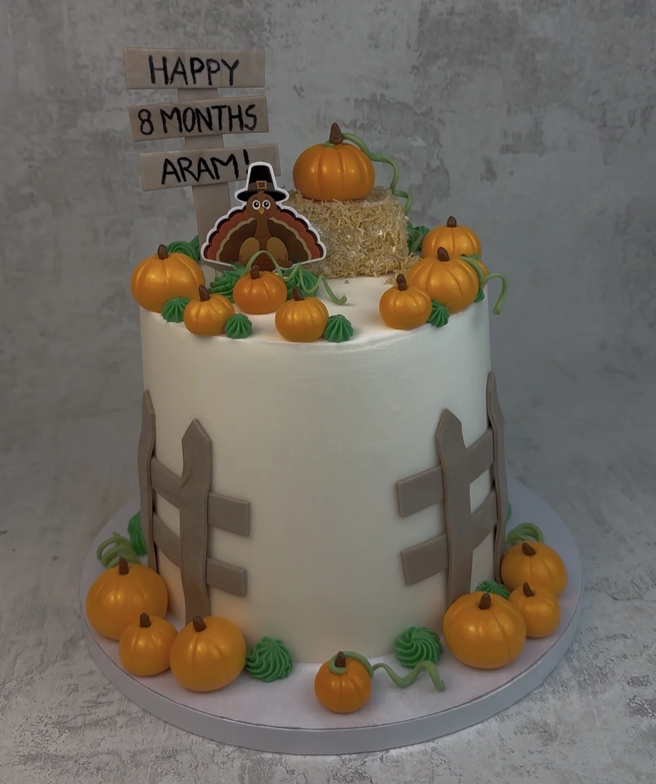 A birthday cake decorated with pumpkins, green swirls, and a fence, featuring a turkey topper and a sign that reads 'Happy 8 Months Aram!'
