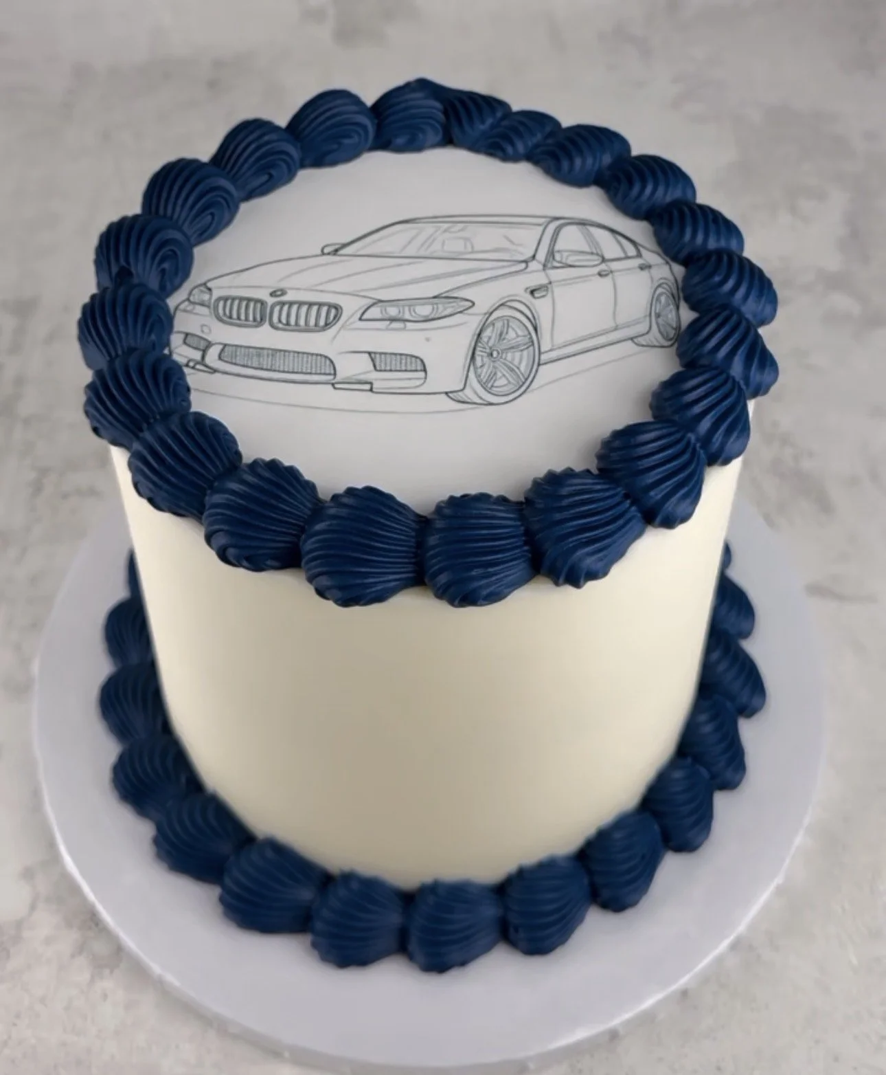 White cake decorated with dark blue piped frosting, featuring a black-and-white drawing of a BMW car on top.