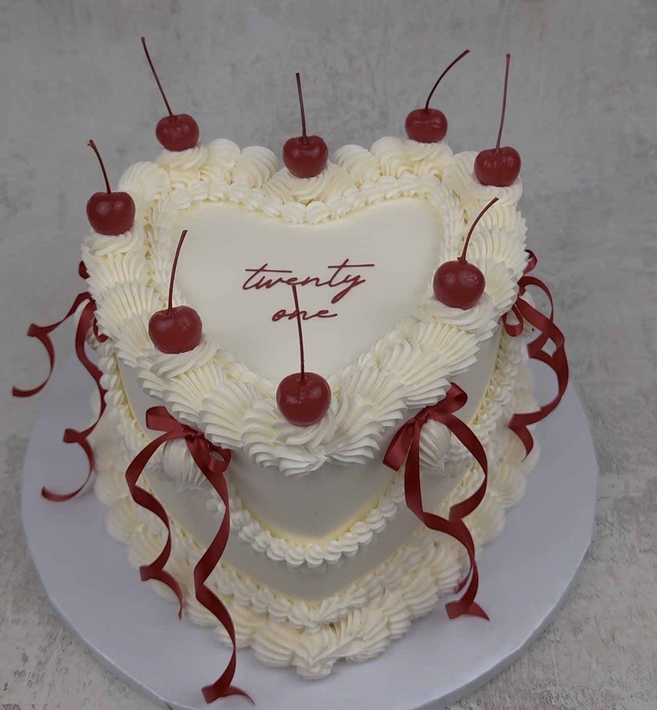 A heart-shaped white birthday cake decorated with whipped cream, cherries, red ribbons, and a message that says 'twenty one'.