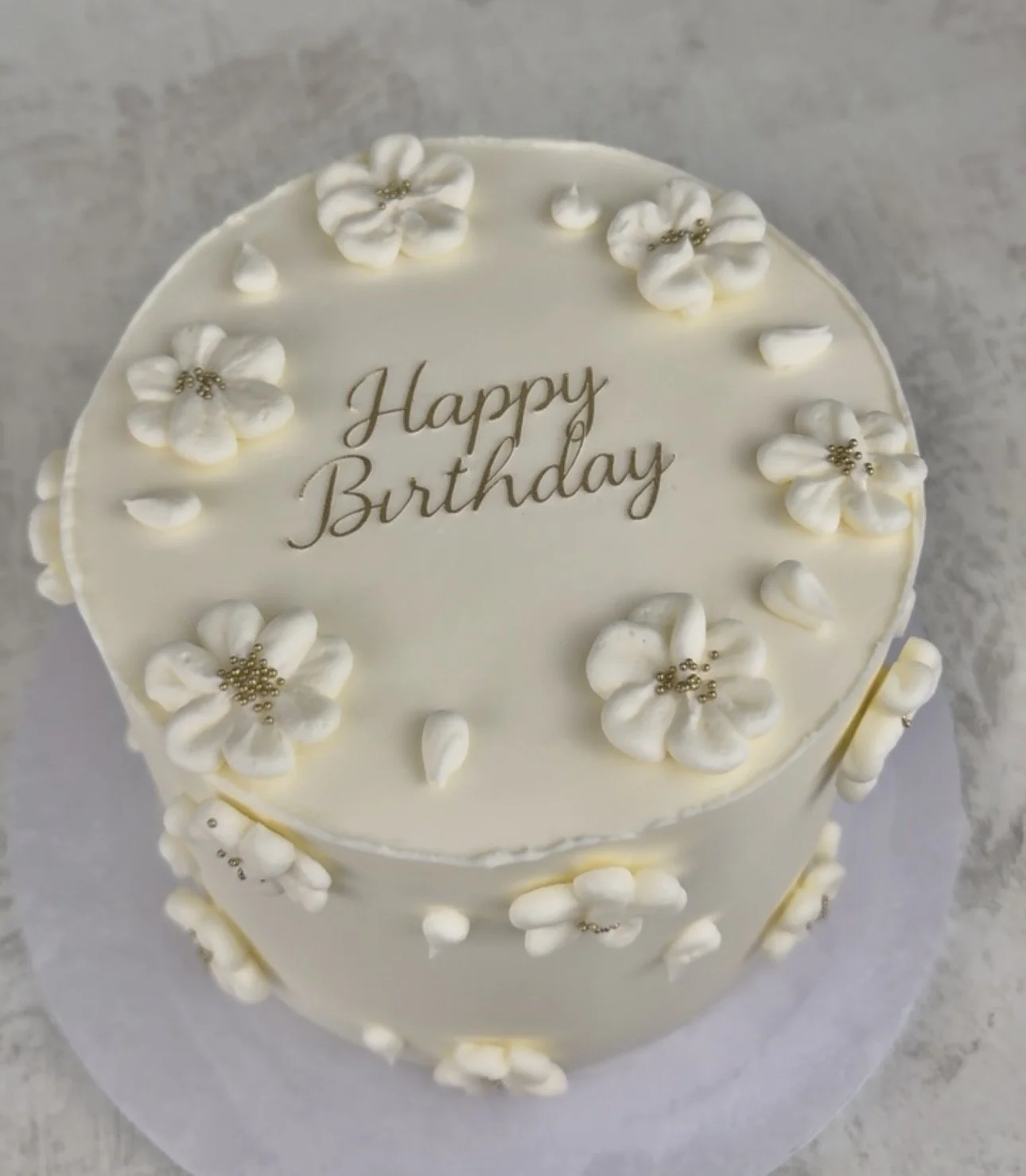 A white birthday cake decorated with flowers and gold beads, with the words 'Happy Birthday' written on top.