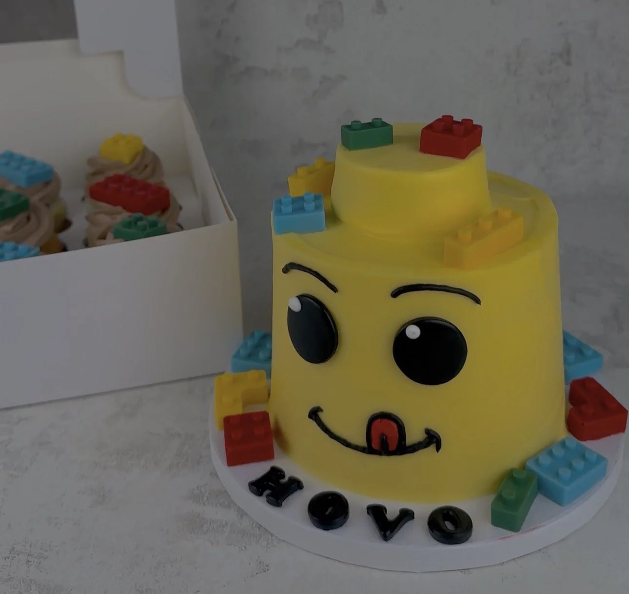 A birthday cake decorated as a cute, smiling Lego character with big black eyes and a tongue sticking out, surrounded by colorful Lego bricks, with 'HOVO' spelled out on the cake board.