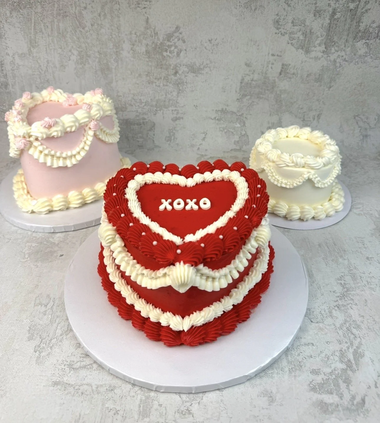 Three decorated cakes, one heart-shaped with red and white icing and the word "XOXO" in the center, and two round cakes with white and pink icing with piped designs, on a gray textured surface.