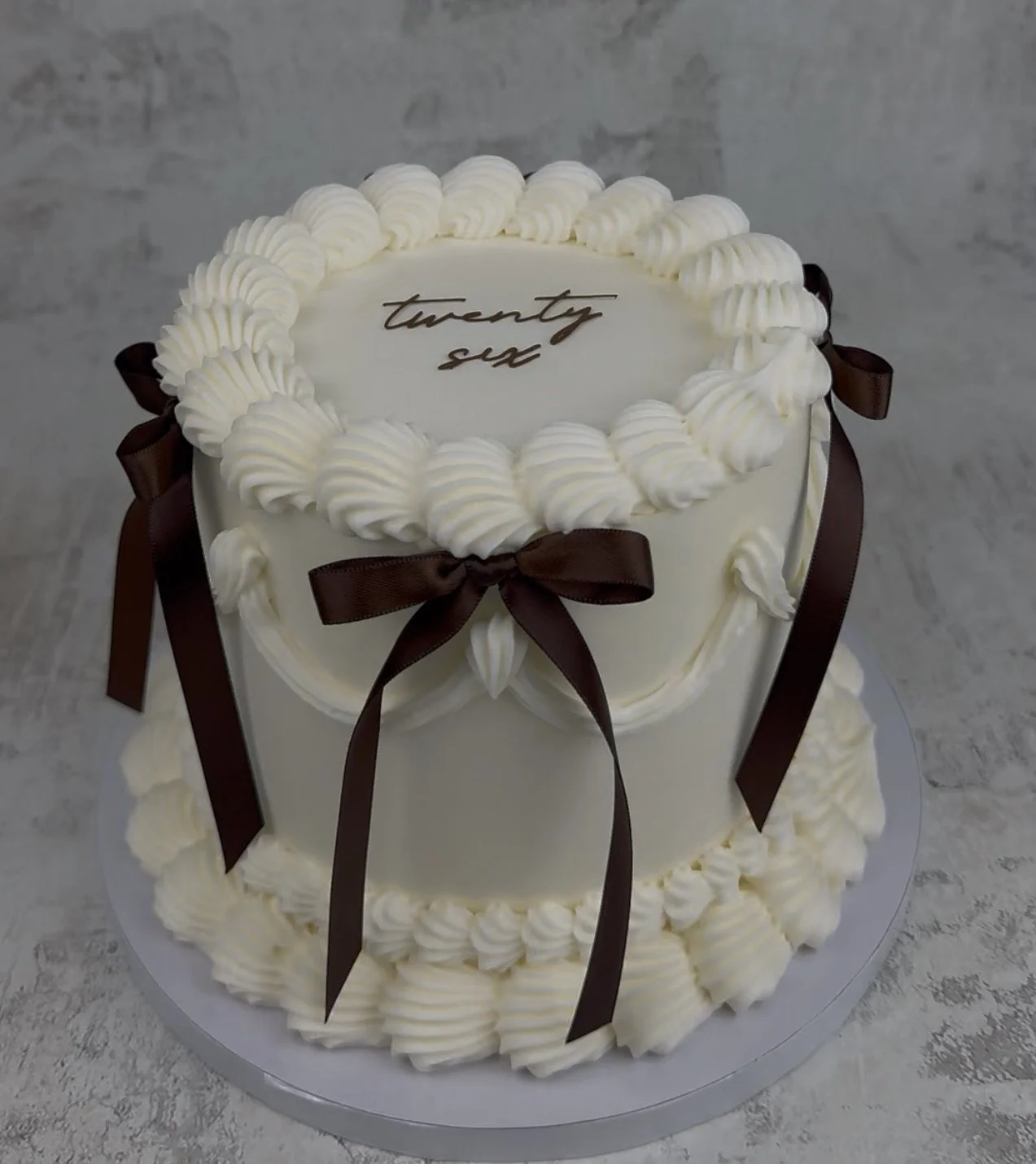 A white birthday cake decorated with white frosting swirls and brown ribbons, with "twenty six" written on top.