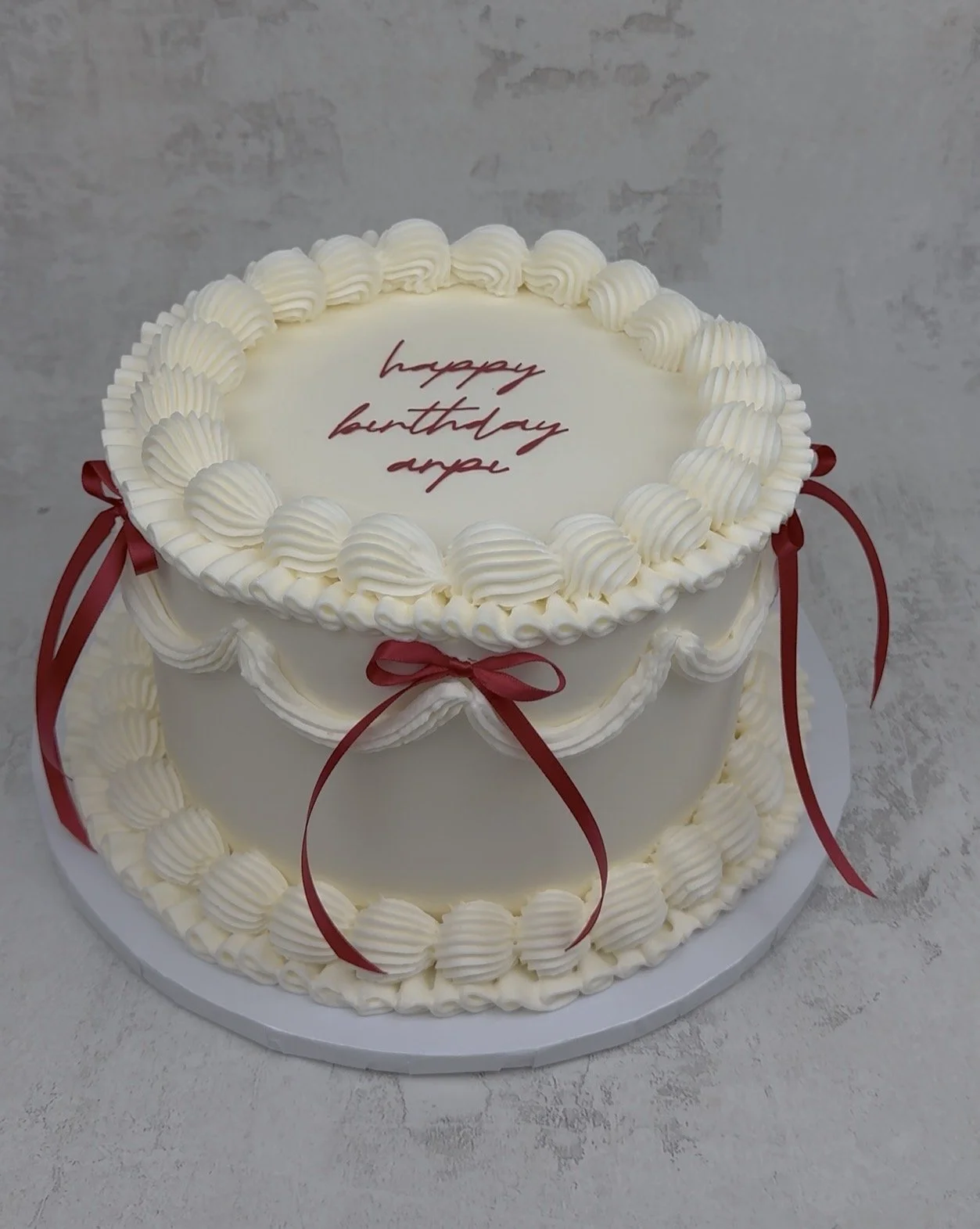 Two-tiered white birthday cake decorated with white piped frosting and red ribbons, with a message written in red icing on top reading 'happy birthday apple'.