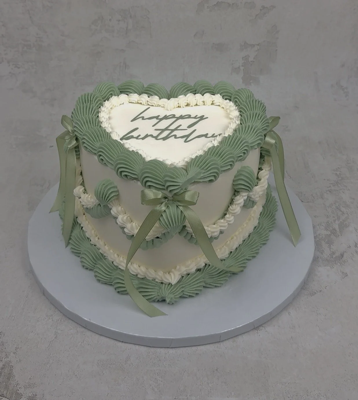 Heart-shaped birthday cake decorated with green and white frosting, green ribbons, and a message that reads 'happy birthday' on top.