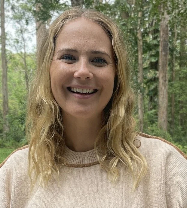 A woman with wavy blonde hair smiling outdoors with trees in the background.