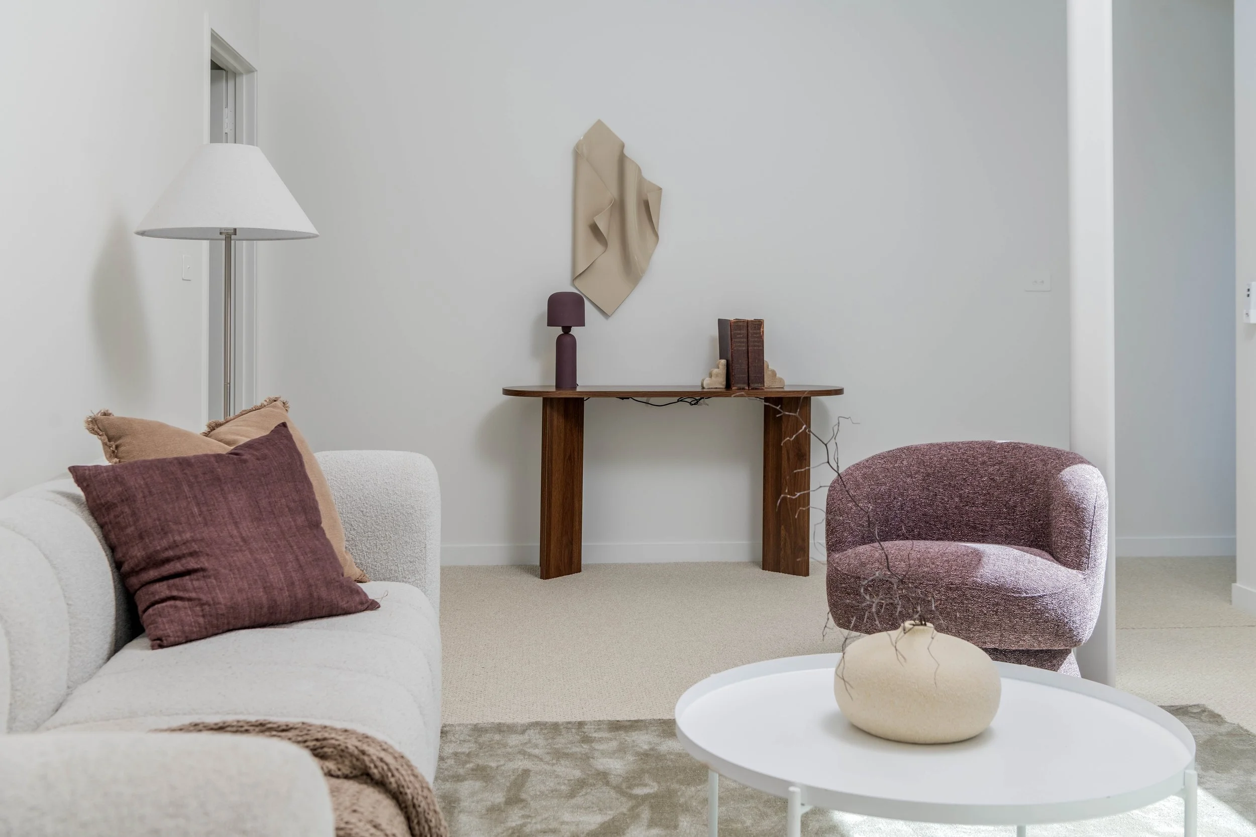 Living room with white sofa, maroon and beige pillows, pink armchair, white round coffee table with a beige vase and branch, wooden console table with books and a purple lamp, white wall with abstract art