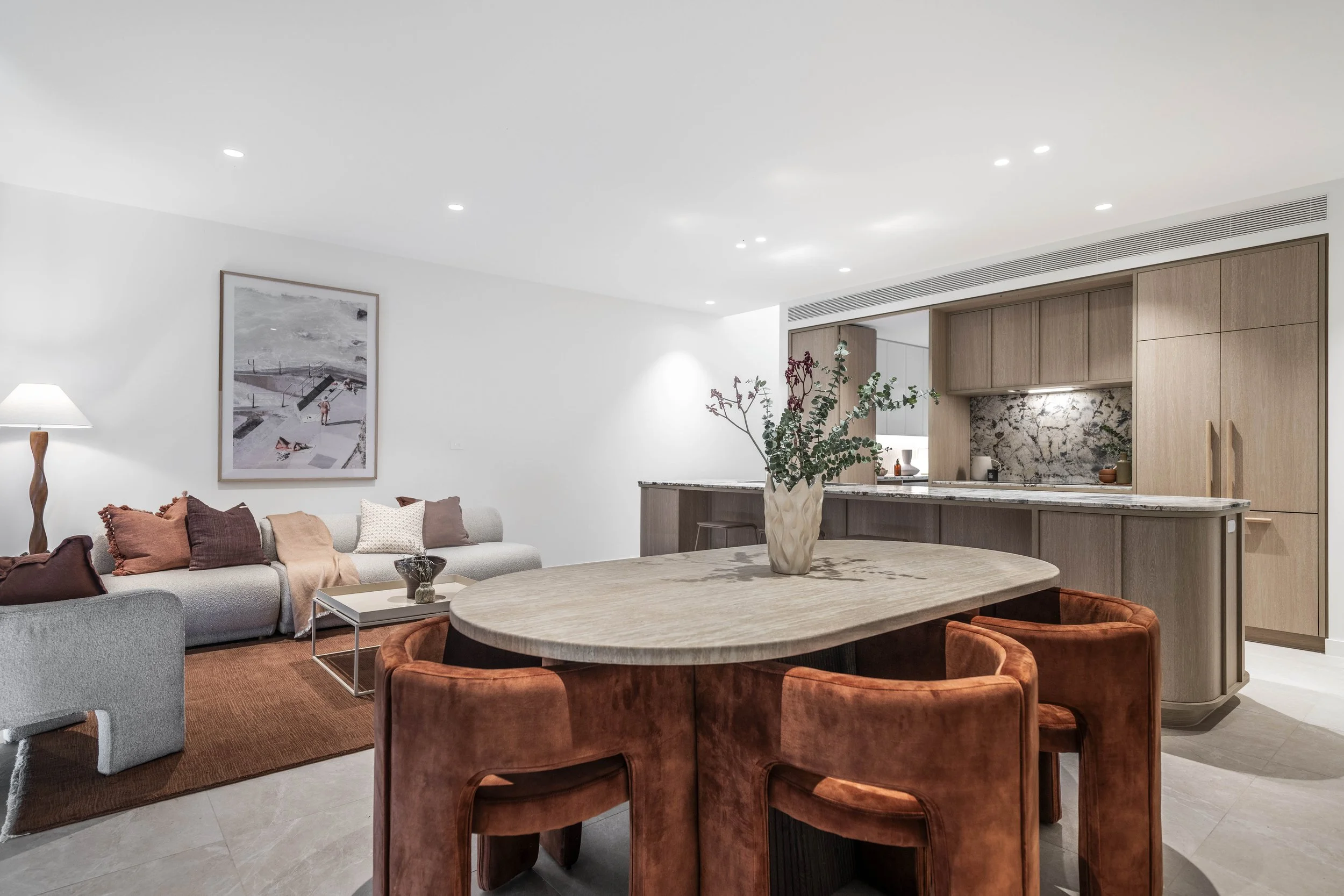 Modern living room with dining area, featuring a wooden oval table, rust-colored chairs, a gray sofa with pillows, a framed art piece, and a kitchen with light wood cabinets and marble backsplash.
