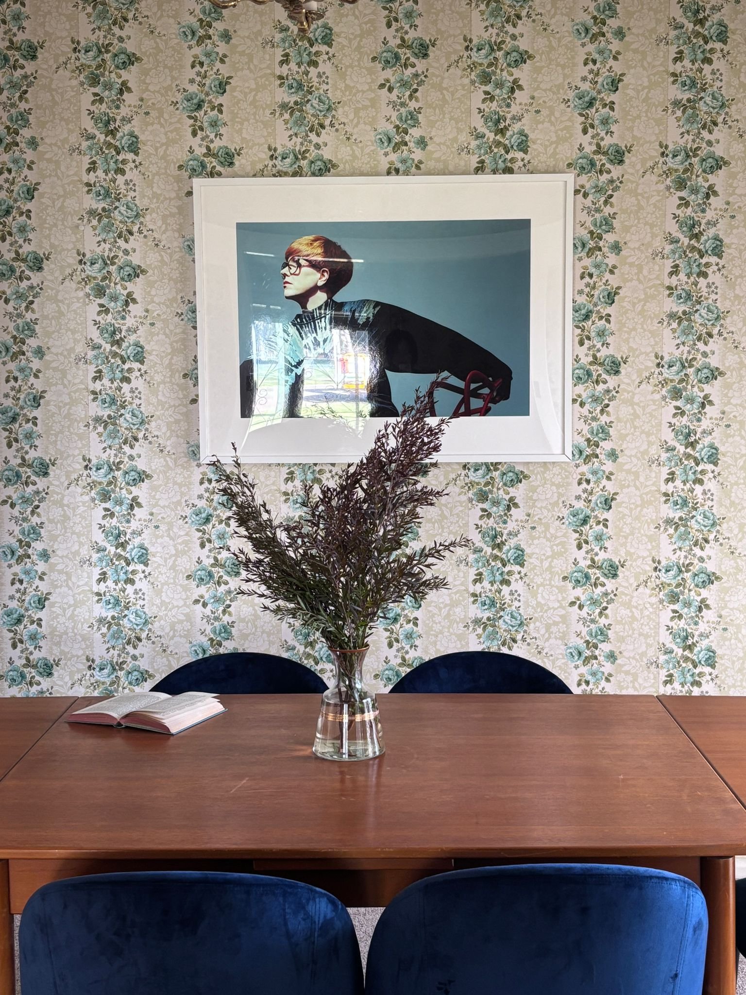 A dining table with a glass vase containing purple-green foliage in the center. An open book resting on the left side of the table. Four blue upholstered chairs, two on each side of the table. Floral wallpaper with light background and blue and green