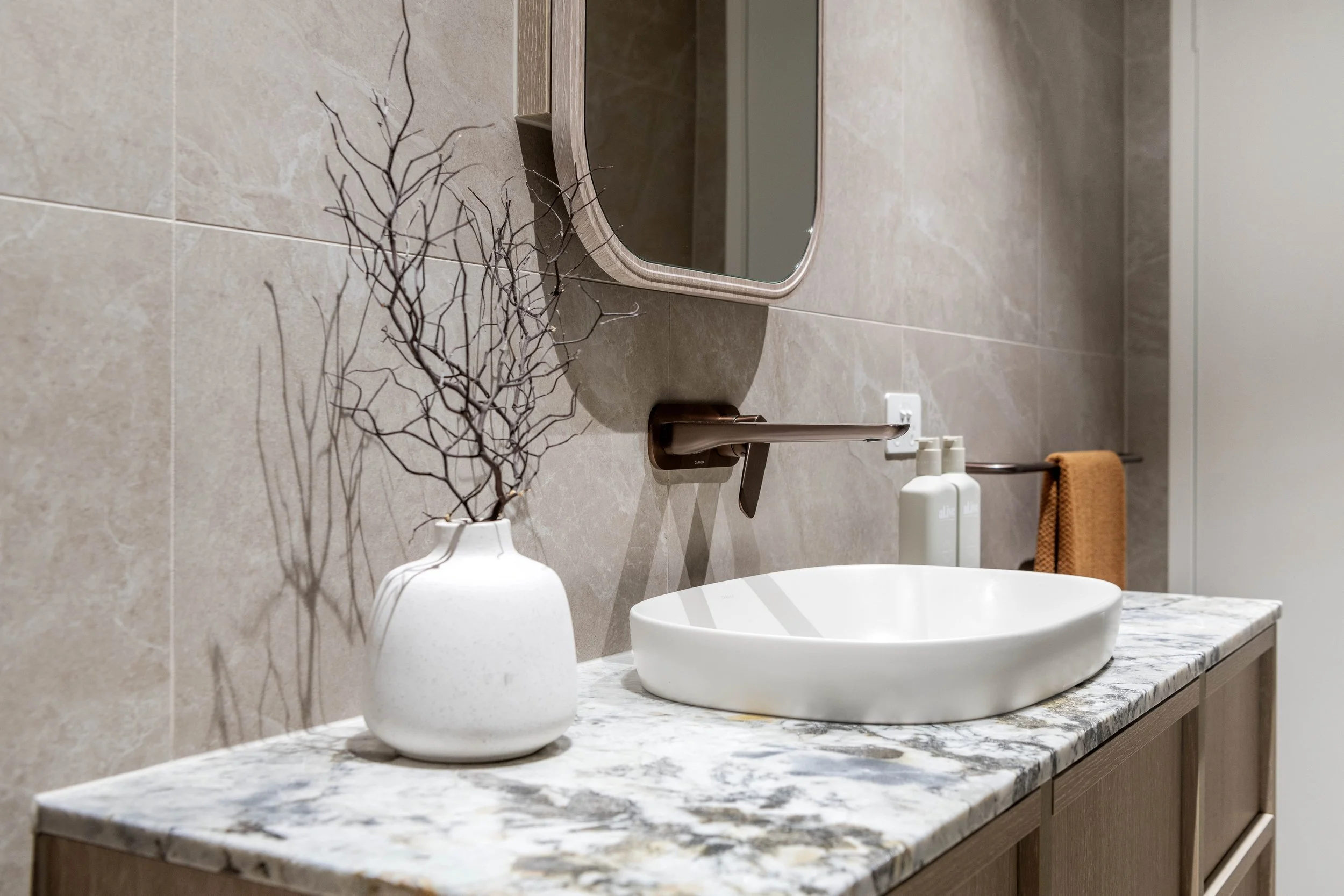 Bathroom sink with a marble countertop, a white vessel sink, a bronze faucet, a mirror, and a white vase with branches, on the right side there are soap dispensers and a towel rack with an orange towel.