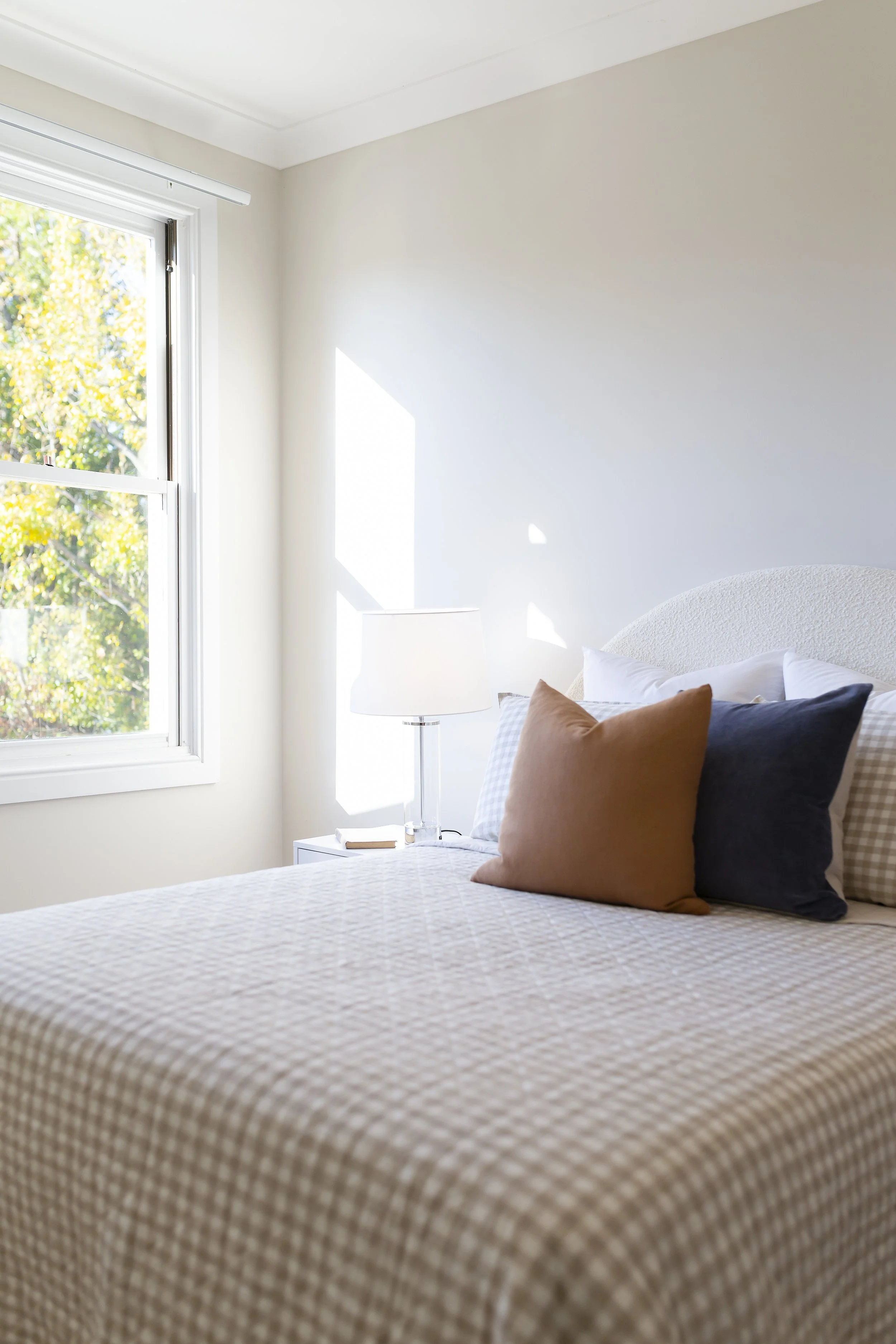 A bedroom with a bed, pillows, a nightstand with a lamp, and a window showing outdoor trees.