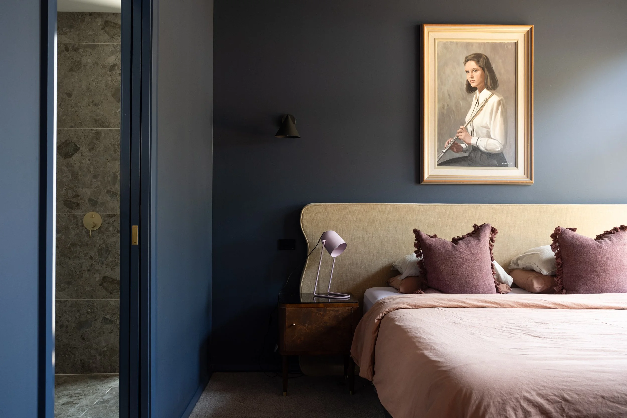 A bedroom with dark walls, a bed with a beige headboard, pink pillows, and a pink bedspread, a small nightstand with a white modern lamp, a framed portrait of a woman with a flute hanging above the bed, and a partially visible doorway to a bathroom w