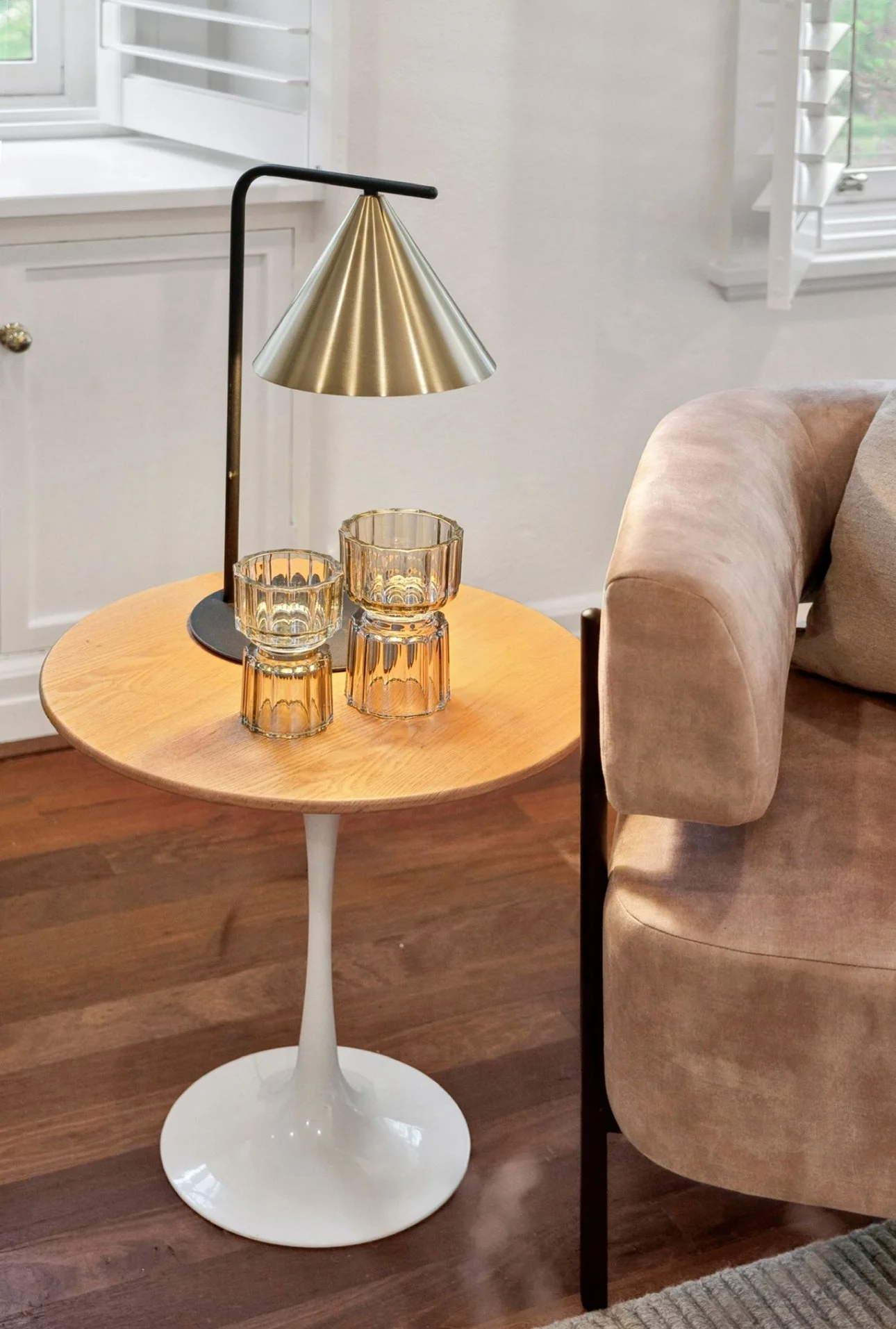 A small round wooden side table with a white pedestal base holding a black and gold modern lamp and two amber glass candle holders, next to a beige upholstered sofa in a well-lit room.
